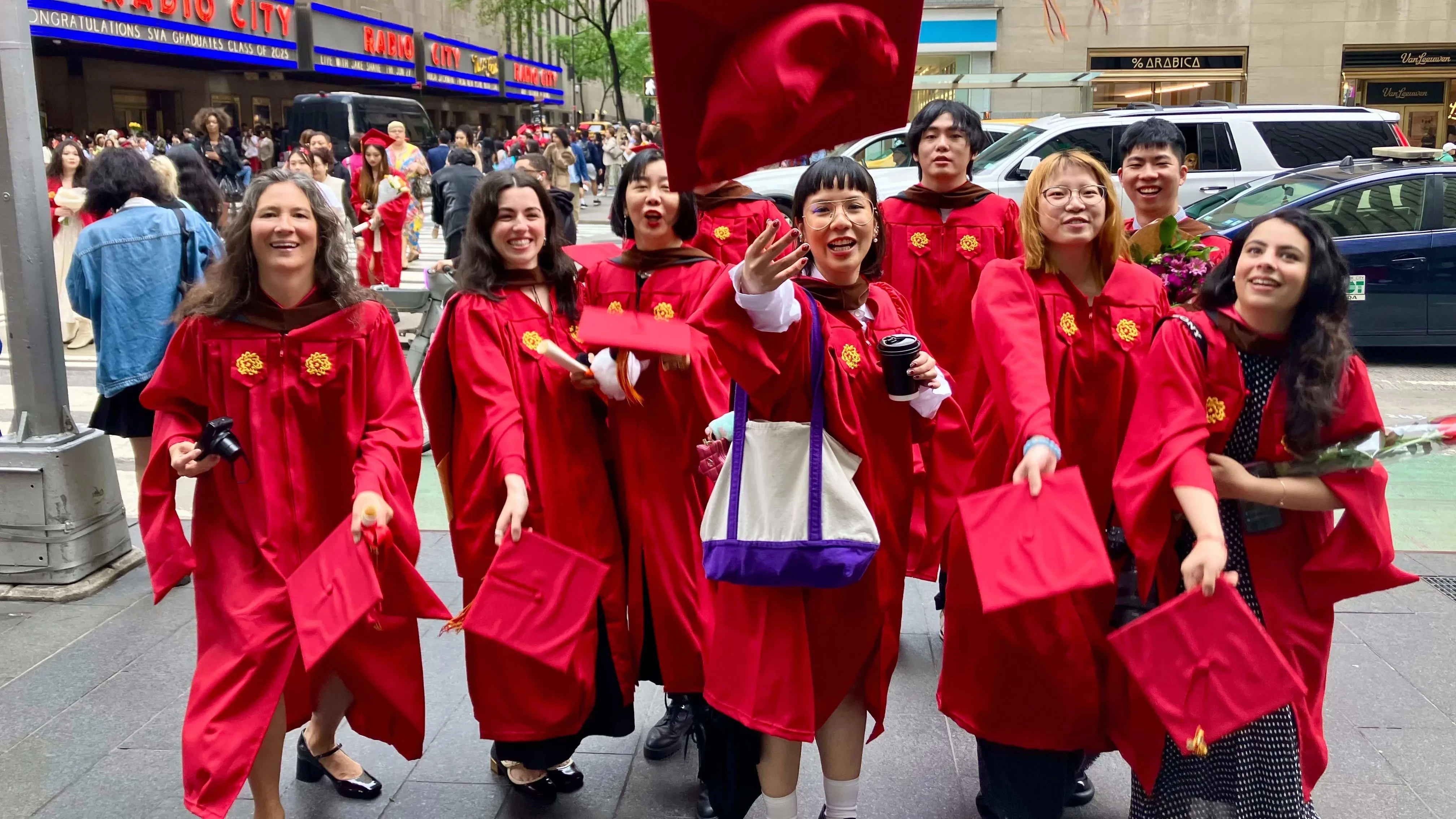 The Masters in Digital Photography graduating class of 2025, in the iconic SVA red graduation gowns, in front of the famed Radio City Music Hall where the commencement ceremony just took place. The students are threwing their caps in excitement. 