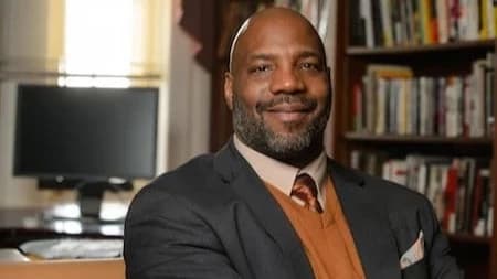 A portrait photograph of writer and academic Jelani Cobb sitting in an office with a desk and bookcase behind him.