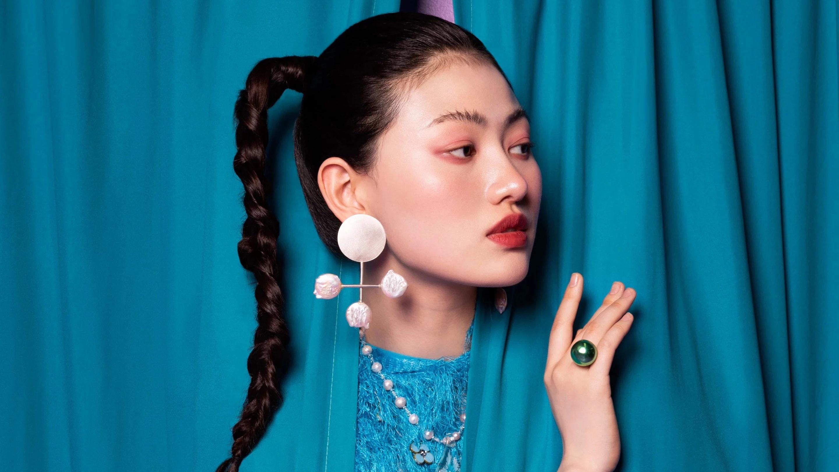 A woman in blue peers out from behind a set of blue curtains. She is wearing a large white earring and a round green ring.