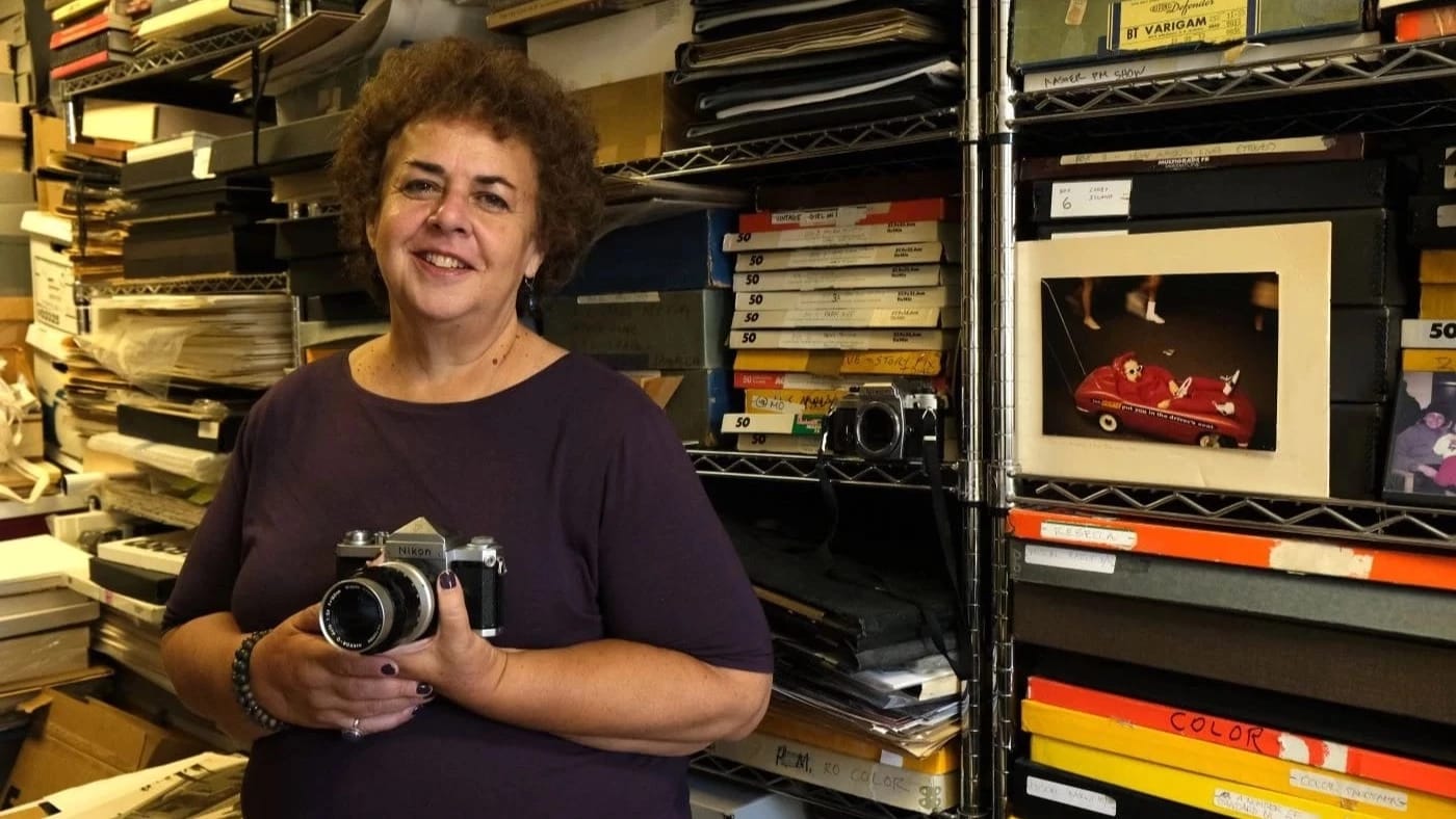 Portrait of Mary Engel taken in front of shelves of photo archives. She is holding a camera and smiling.