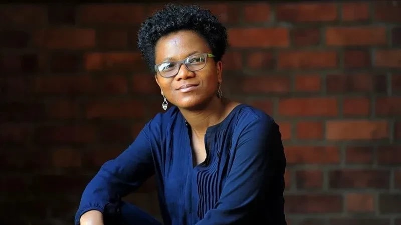 a person in a blue sweater sits in front of a brick wall and smiles at the camera 
