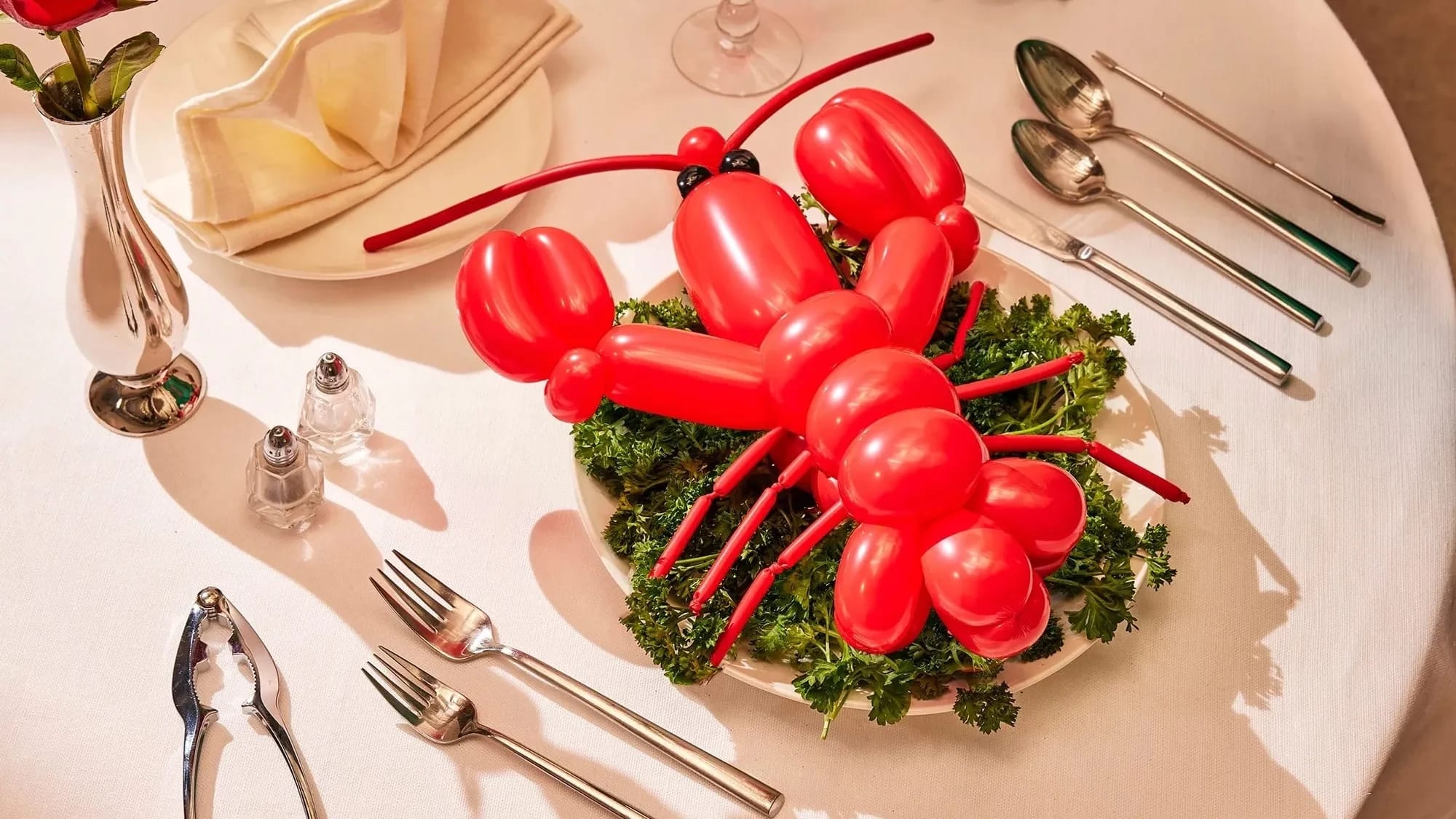 A photograph of a lobster shaped red balloon sitting on top of a bed of kale leaves on a dinner plate. Surrounding the scene is a dinner table fully set up with cutlery,  a dinner napkin folded into a fan, and a single rose in a tin vase.