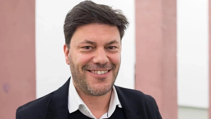 a smiling person with short brown hair and a black blazer on stands in front of a pink and white building exterior 