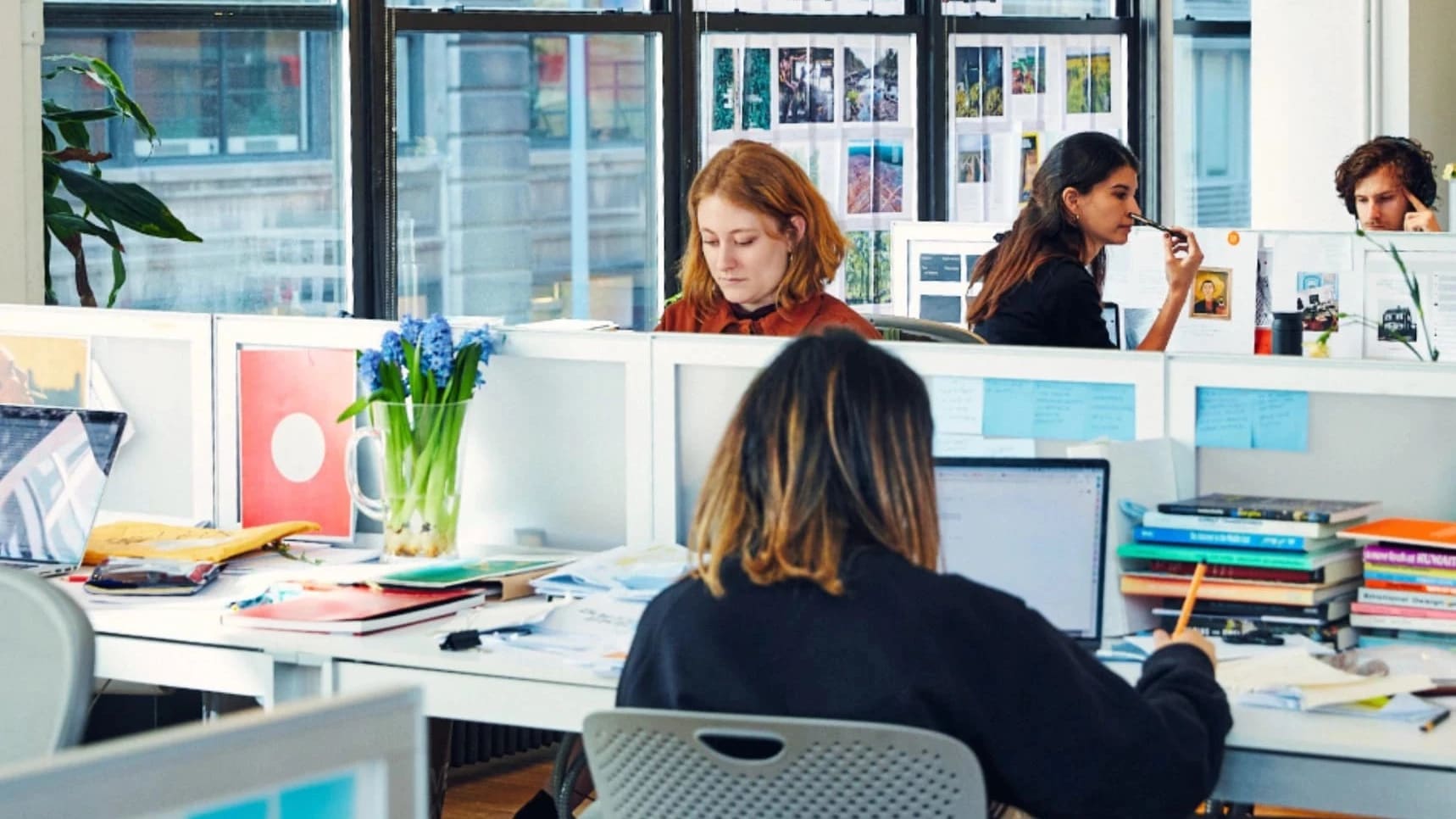 A person with orange hair sits at a row of desks working while other people in the room also work  