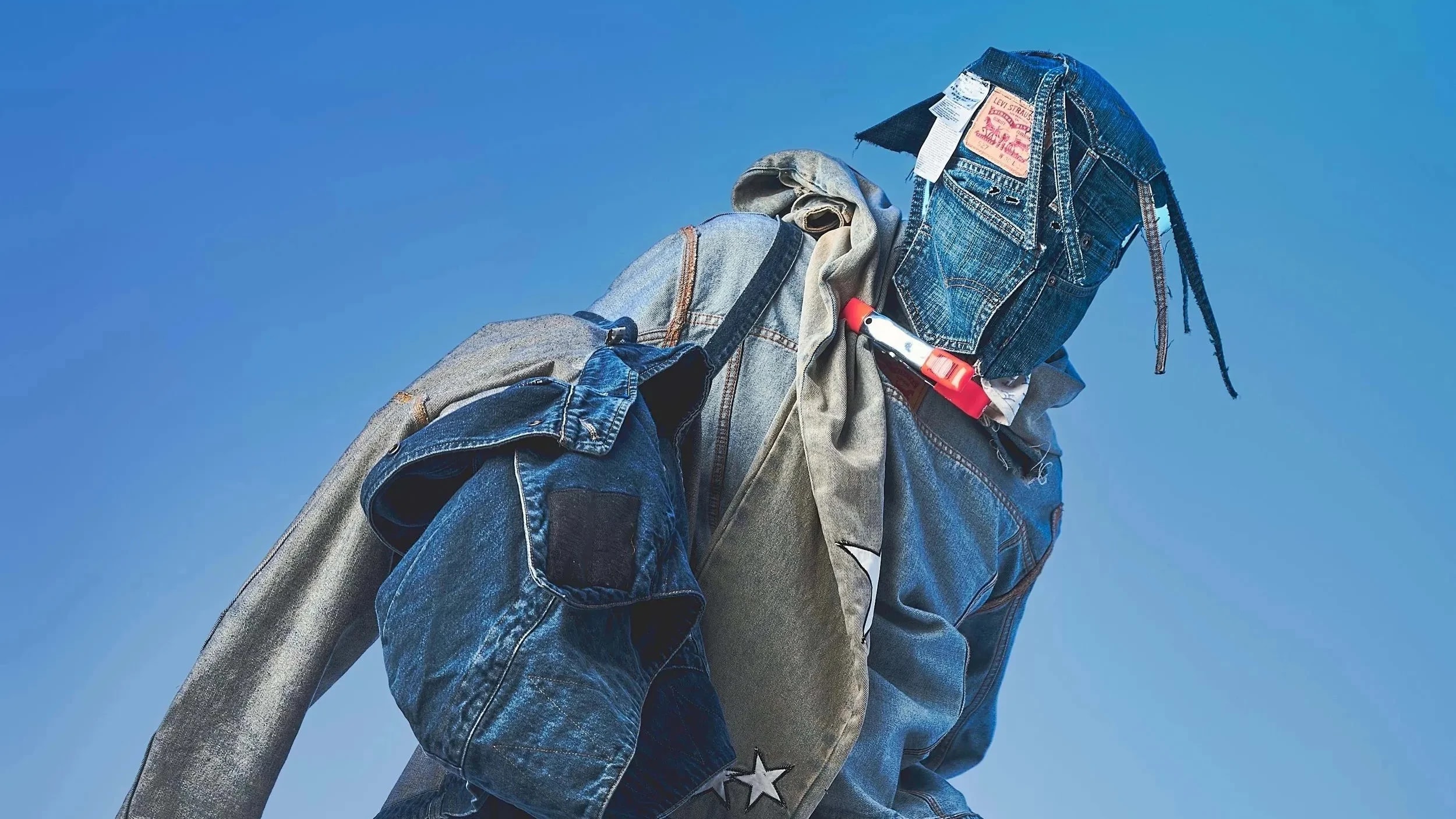 A studio fashion portrait of a model covered in an all-denim blue outfit and wearing a mask made of denim. The model stands in front of a blue background.