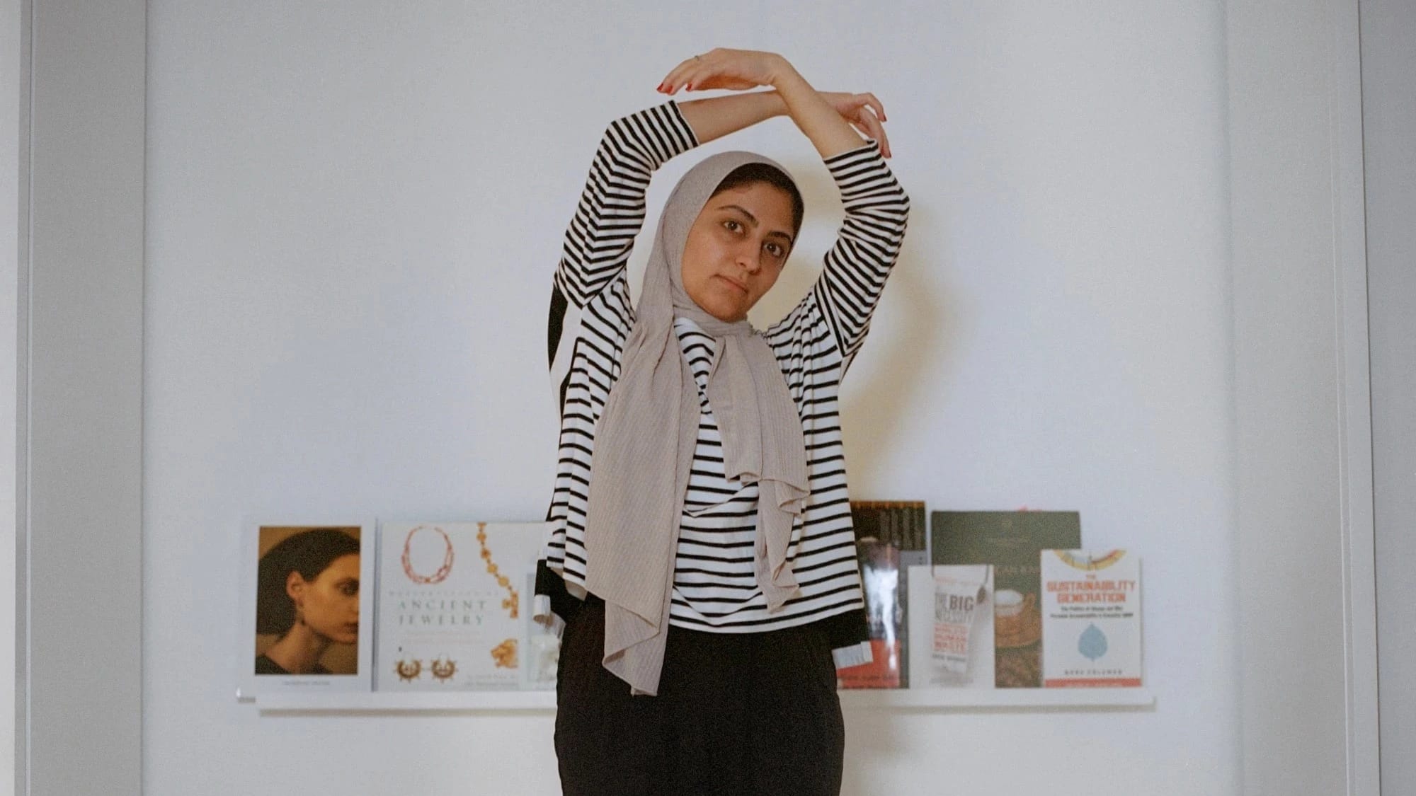 A woman stands on a white bed with her arms circled over her head. She is wearing a headscarf, striped shirt and black pants.