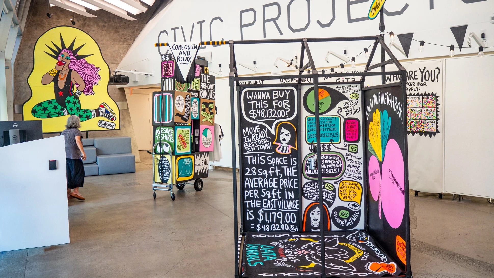 Interactive data visualization installation in a modern gallery space featuring colorful, street art-inspired panels with housing cost data. The central black panel displays NYC housing statistics including '$48,132.00' and text about East Village rental prices per square foot. Surrounding panels feature vibrant graphics with pink, yellow, and green elements. A person in casual clothing examines the display while standing on polished concrete floors under industrial ceiling architecture.