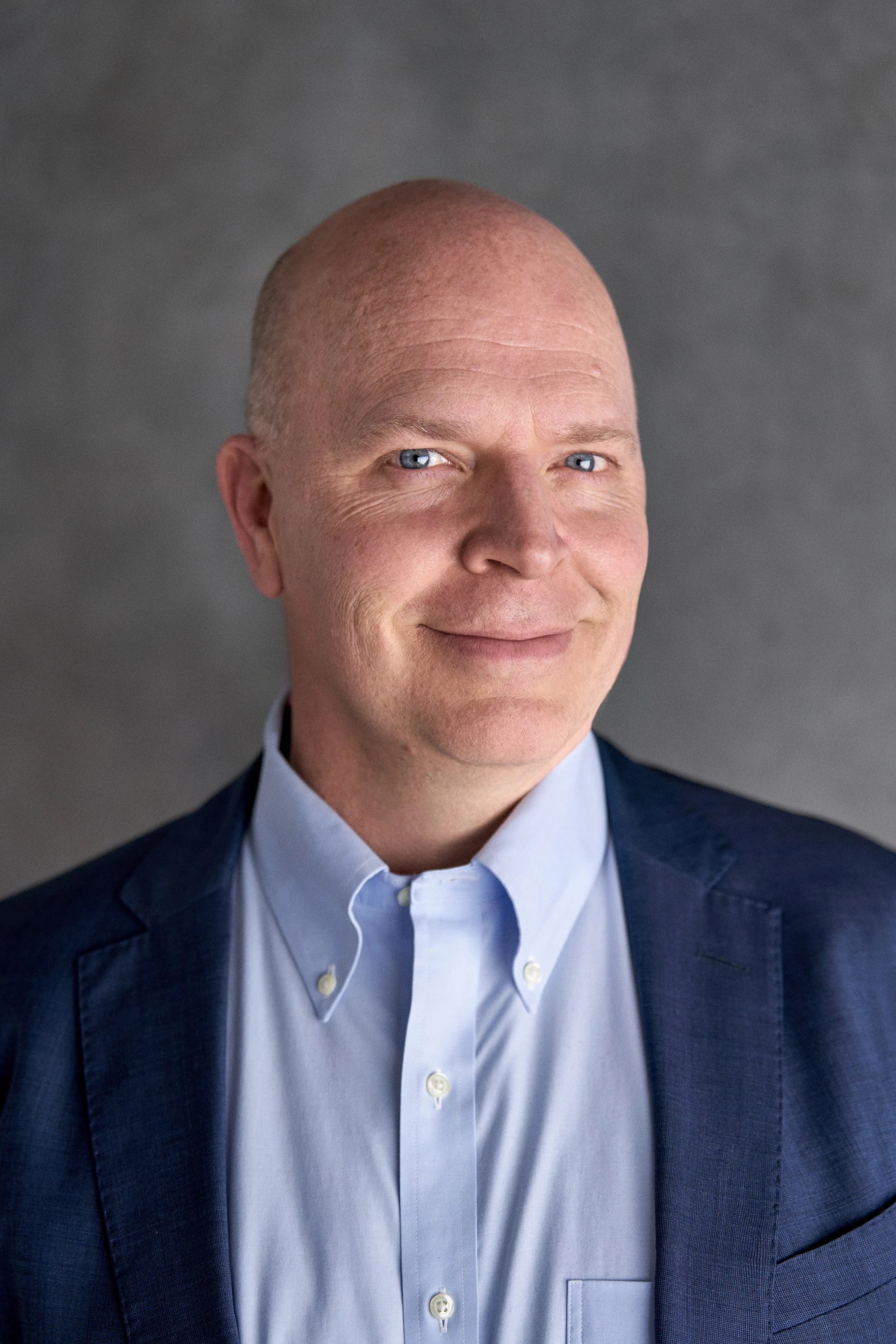 A middle-aged man with light skin and blue eyes, wearing a light blue button-down shirt and a navy blazer. He is smiling slightly and looking directly at the camera. The background is a neutral gray, creating a professional and approachable tone.