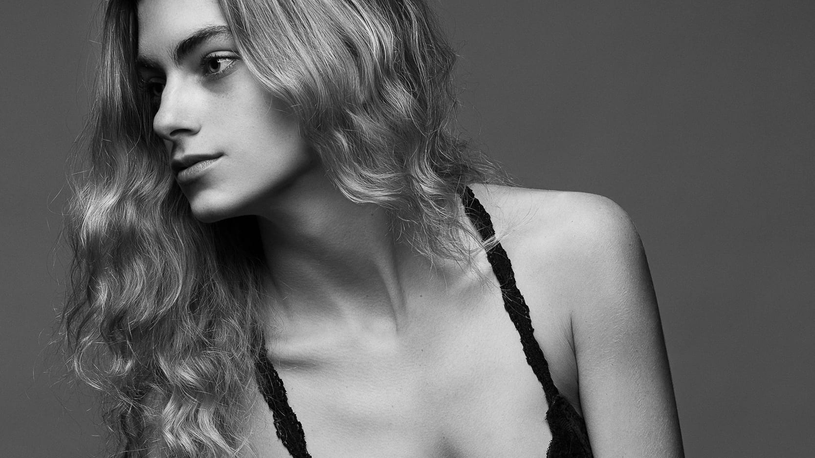 A black-and-white studio photograph of a woman sitting on a black surface. She is leaning slightly to the left and looking to the left. She is wearing a black sports bra and high waisted black underwear. She has long, wavy hair.