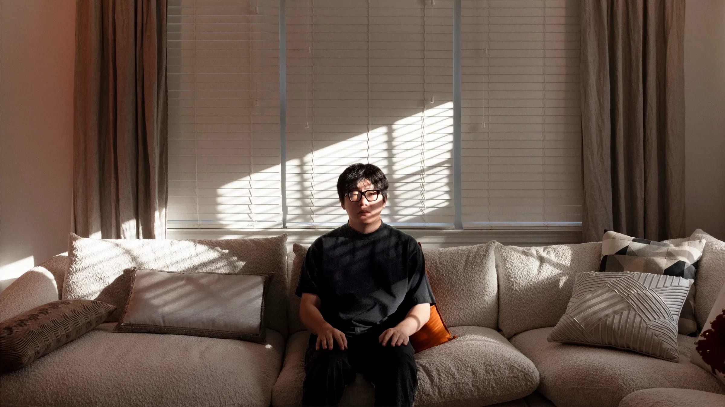 In this color photograph, the artist sits at a couch while looking into the camera. Behind him are drawn curtains.