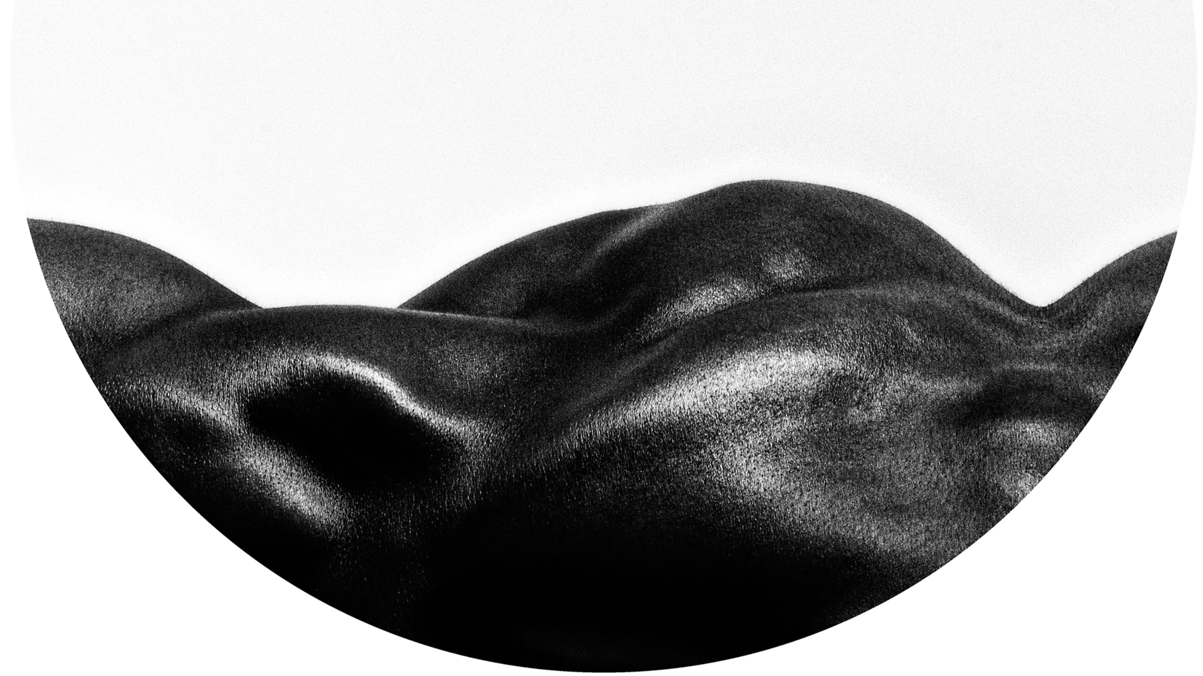 An abstract photograph of part of a black person’s body laying down and photographed from the side. It is against a off-white background. The whole image is framed as a circle vignette. The rest of the image outside the circle is plain white.