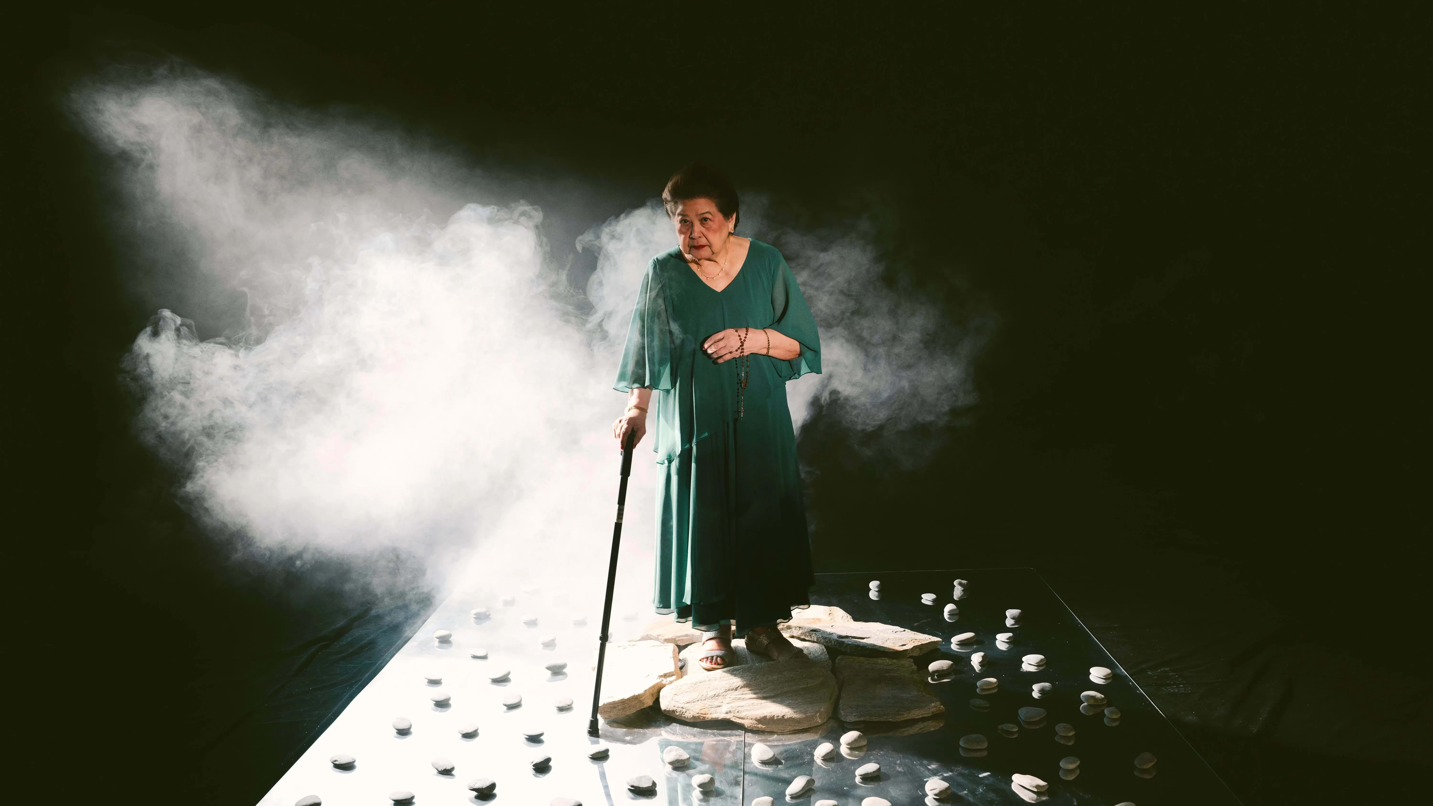 A still from a color video: An elderly woman in a teal dress stands on a cluster of flat stones, holding a cane in her right hand and a rosary in her left. She is surrounded by smoke and placed on a glossy black platform scattered with smooth white stones. The background is dark, with light focused on the figure from the left.