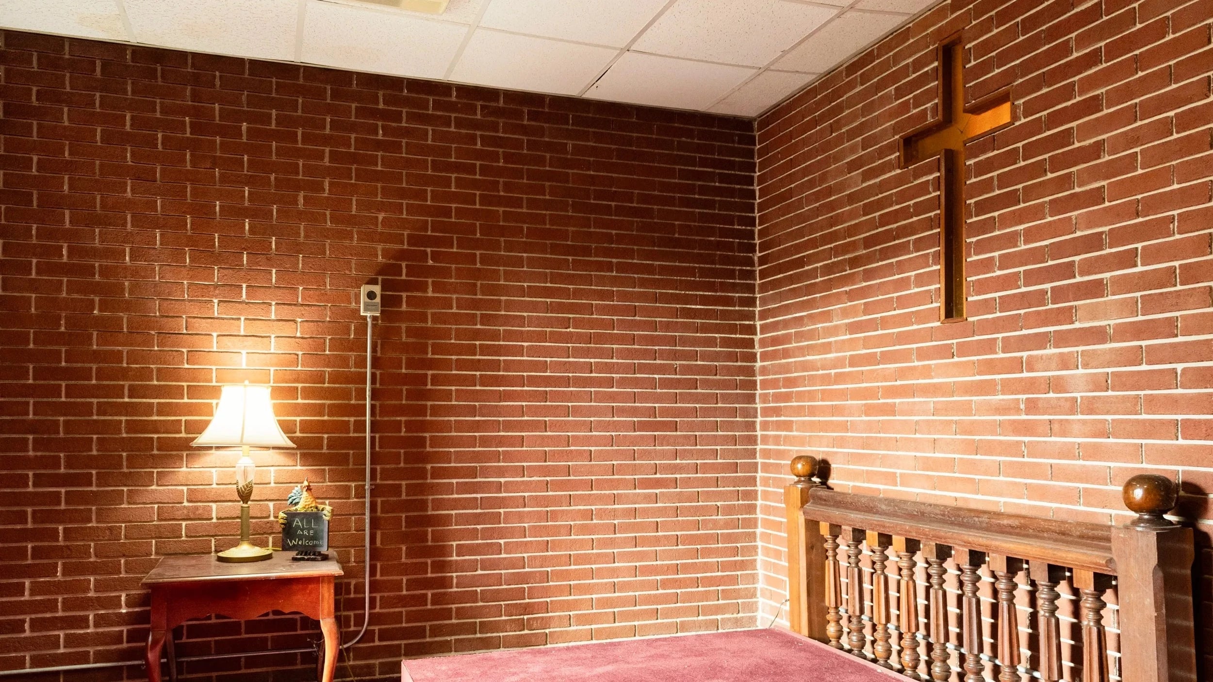 A small prayer room with an altar, side table, and lamp. A cross is embedded in the brick wall.