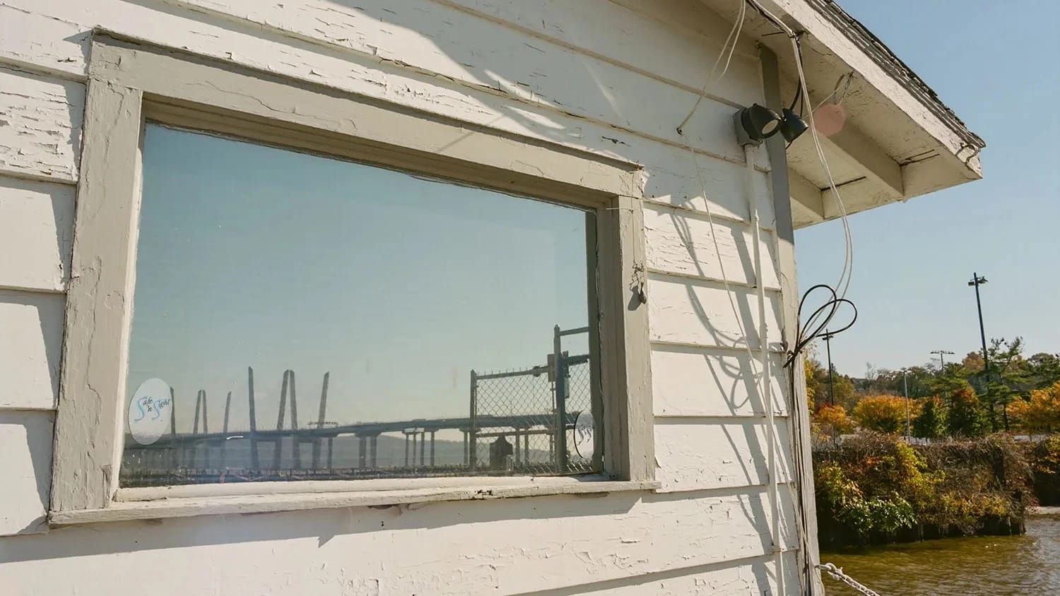 The window of a white shed frames a bridge stretching across a river with autumnal foliage in the background.