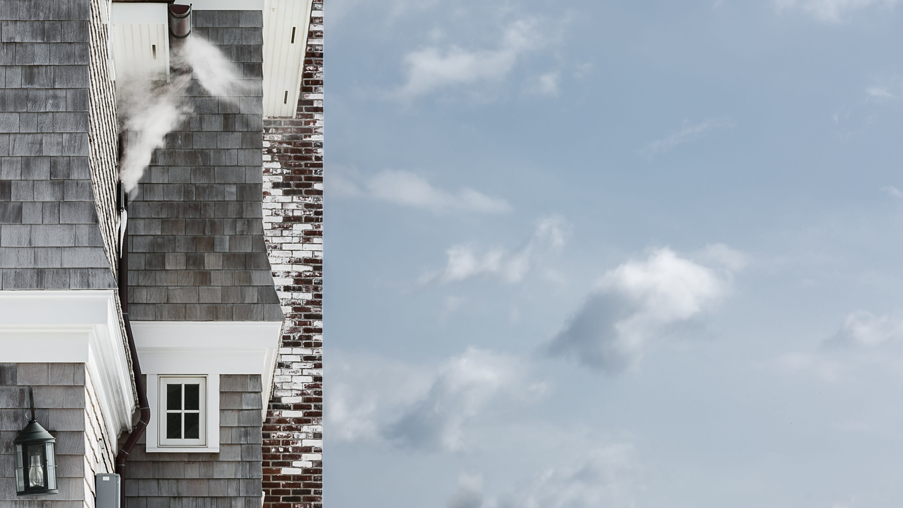  A photograph of a blue sky with clouds. In the left ⅓ of the frame is part of a brown shingled house and its bricked, partially-white painted chimney. From one of the windows, steam is billowing up towards a gutter.