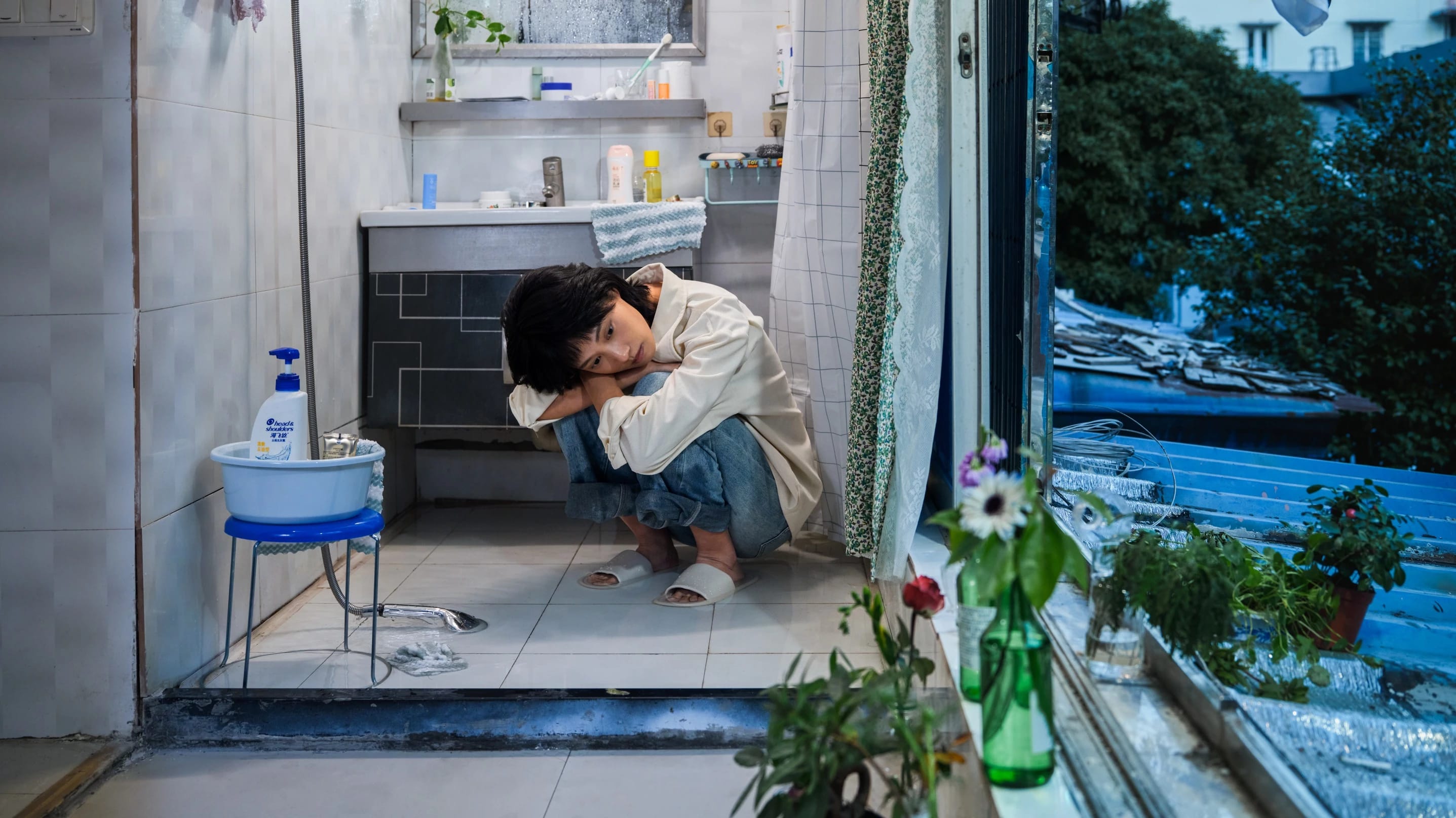 A color photography by Shaohan Fang entitled En Route. In a dim bathroom at dusk, the girl crouches on a wet floor, staring at the window. Water pools around her feet, she appears visibly overwhelmed.