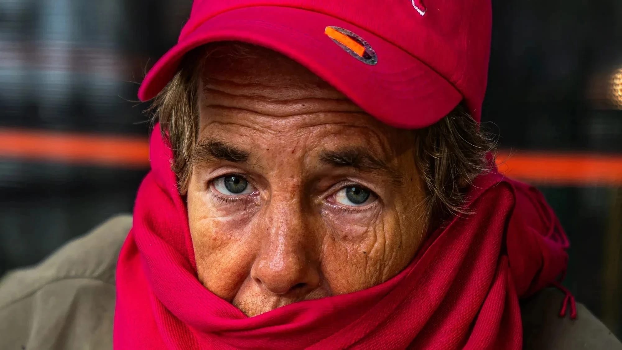 A portrait of an elder person with blue eyes and wrinkles on their face. They wear a red hat, red scarf and green coat.