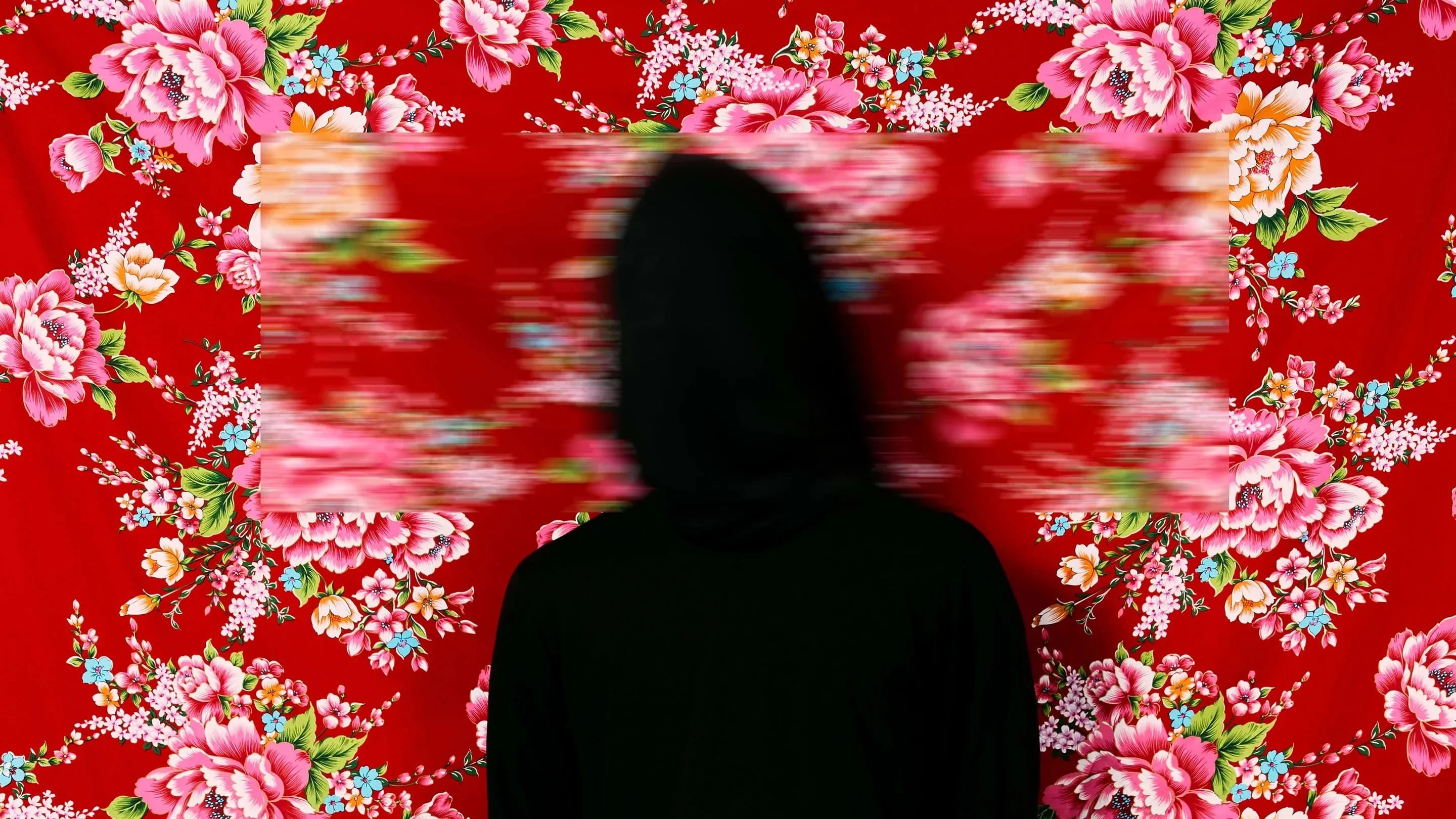 A color photograph featuring a figure wrapped in black from head to toe standing facing the camera against a red Hakka floral fabric background, which is suspended and reveals a faint dark wooden floor below. Three blurred rectangles cover the head, hands, and feet.