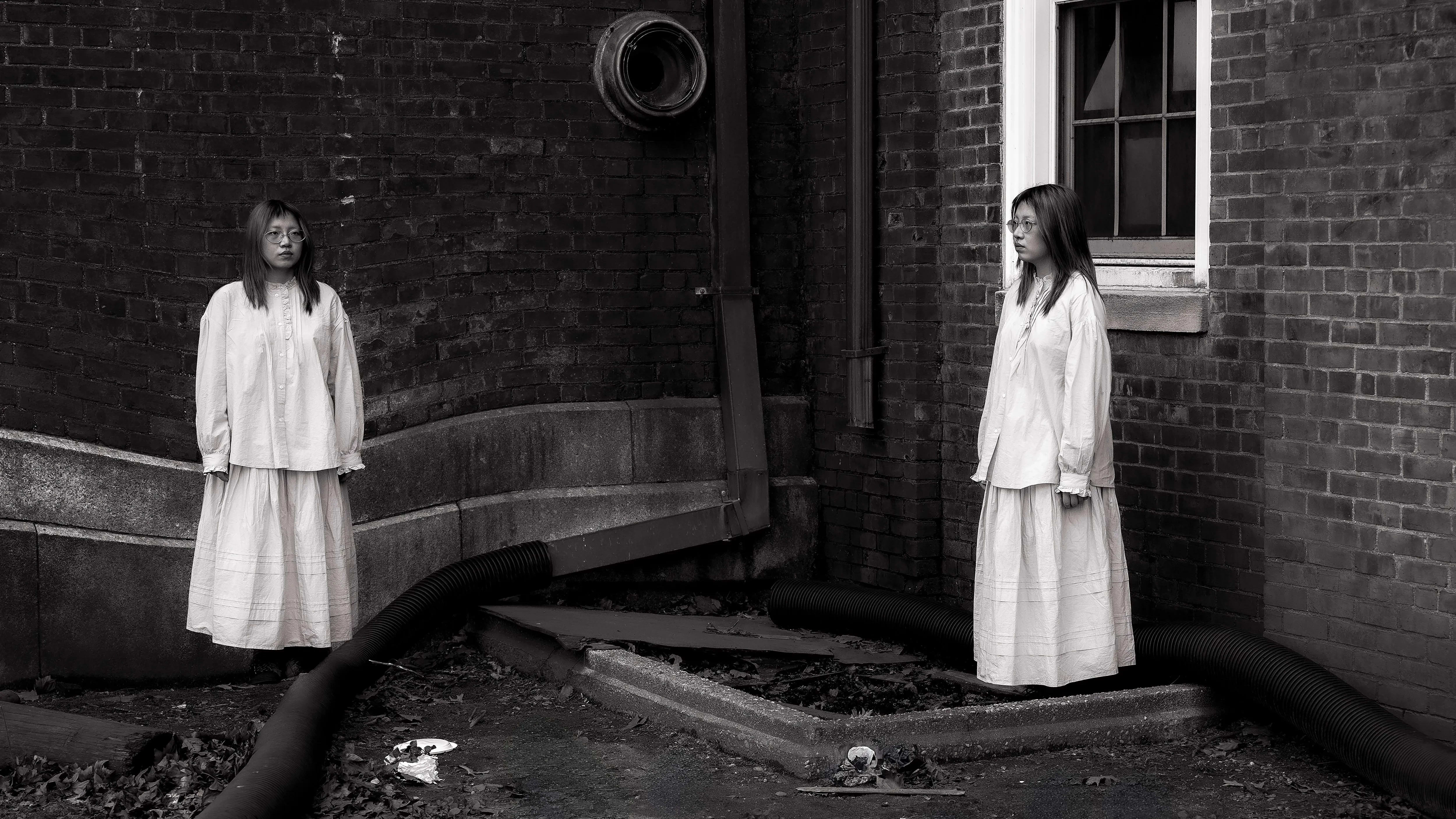 Black and white photograph of a girl duplicated in the frame, standing at two corners of a brick building. Both wear the same long dress, facing each other across a barren, debris-littered ground with industrial pipes.