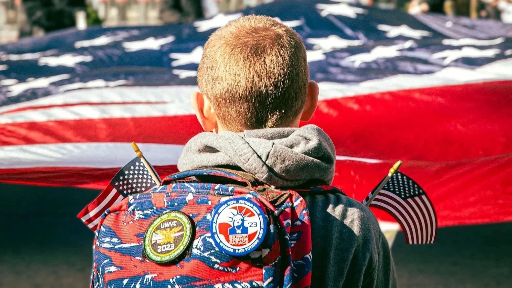 A kid faces away from the camera to look at a large american flag. He has patches on his backpack and holds two small american flags.
