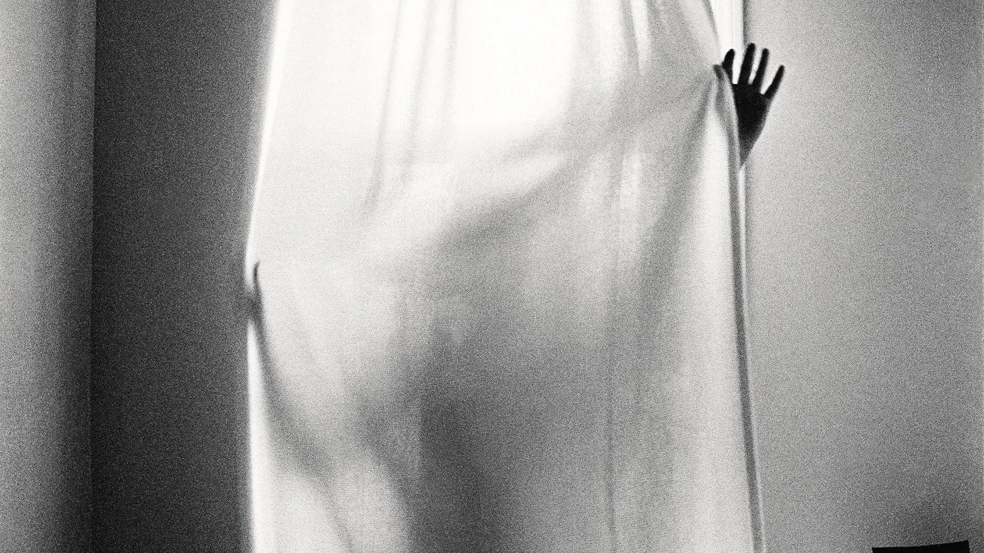 A black and white photo of someone behind a window curtain. The light coming from the window outlines the person's body.