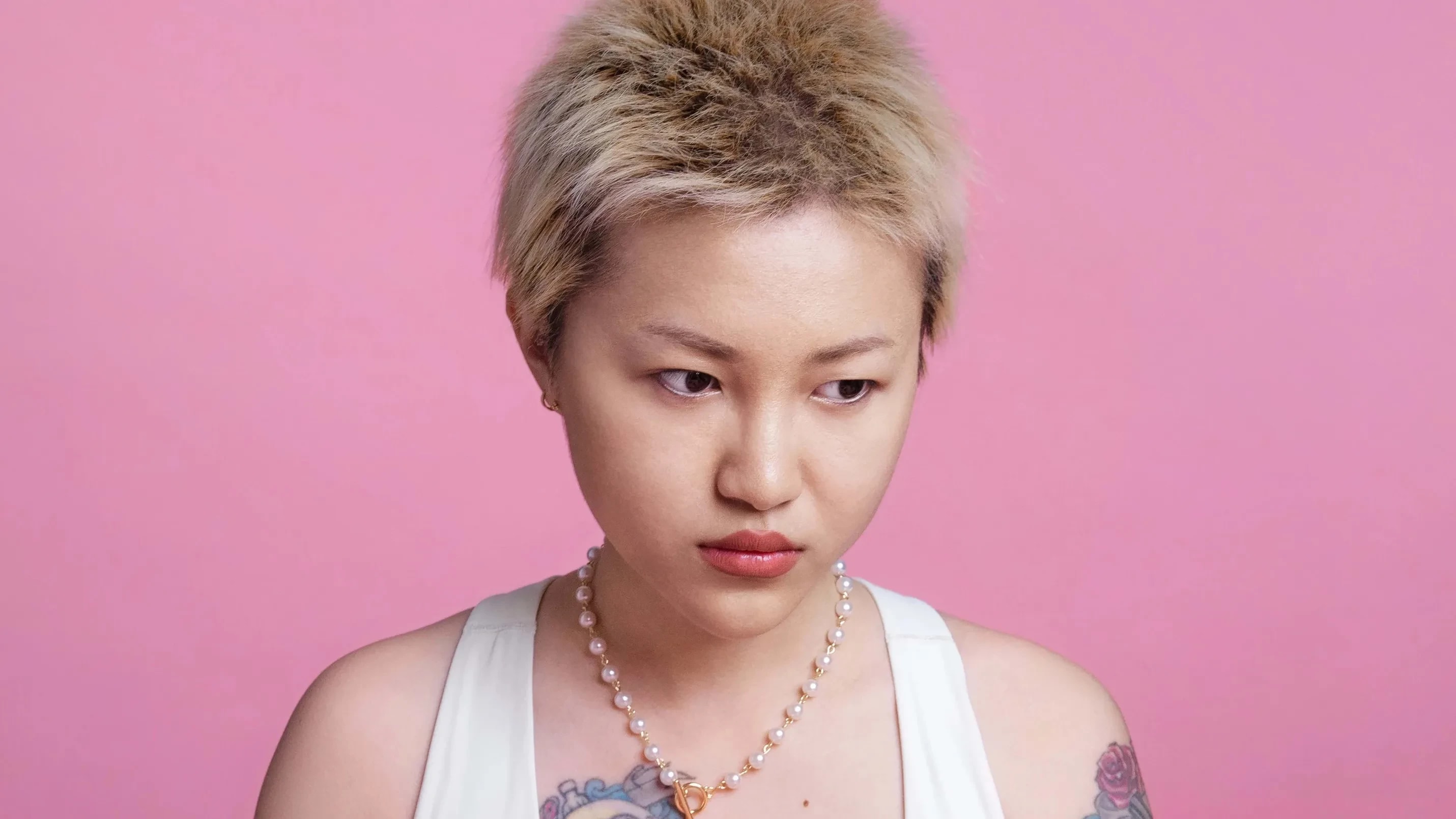 In this color photograph: On a pink background, a half-length portrait of a gold-haired young Asian woman wearing a white sports bra, sitting. She has large colorful tattoos on her right chest and left arm. On the pink table in front of her are three red tomatoes. She is holding a smaller dark- colored tomato and is looking away from the camera, appearing thoughtful.