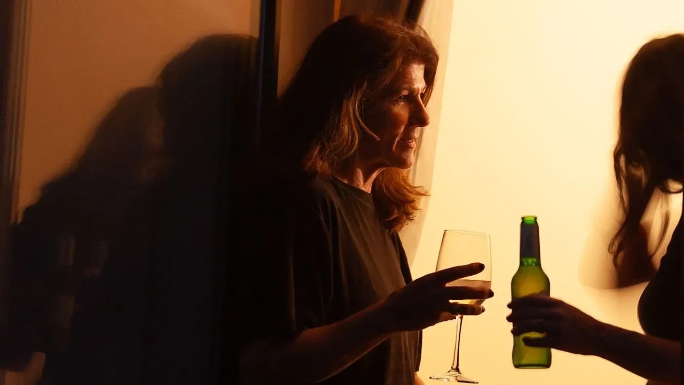 A color photograph of two women by a large open window at nighttime. They are in conversation and one woman holds a wine glass while the other holds a beer bottle.