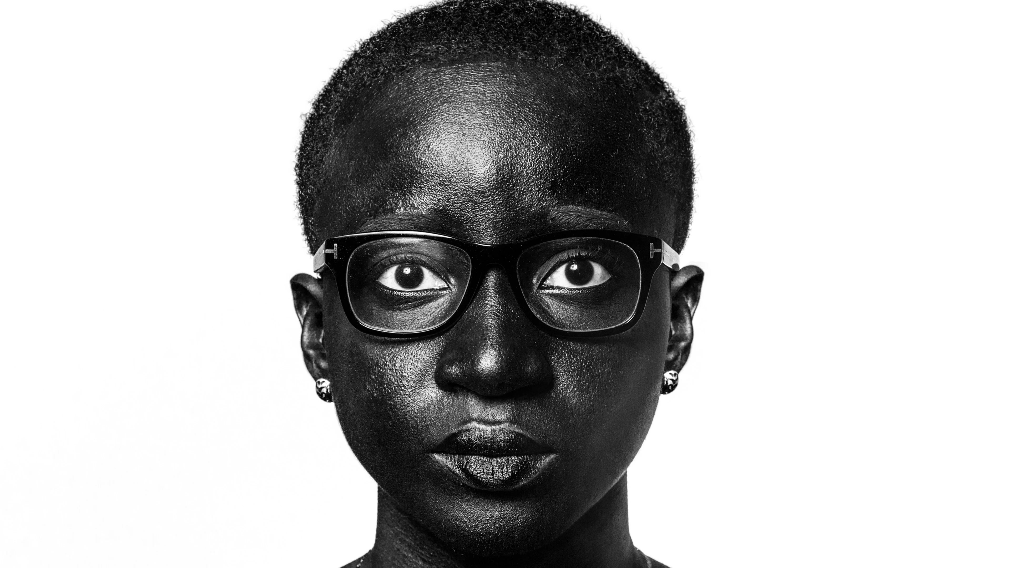 A studio portrait of a black woman from the shoulders up. She is wearing black glasses, a silver necklace, and silver stud earrings. She is staring at the camera with a neutral expression. She has very short, close-cropped hair. The background is white.