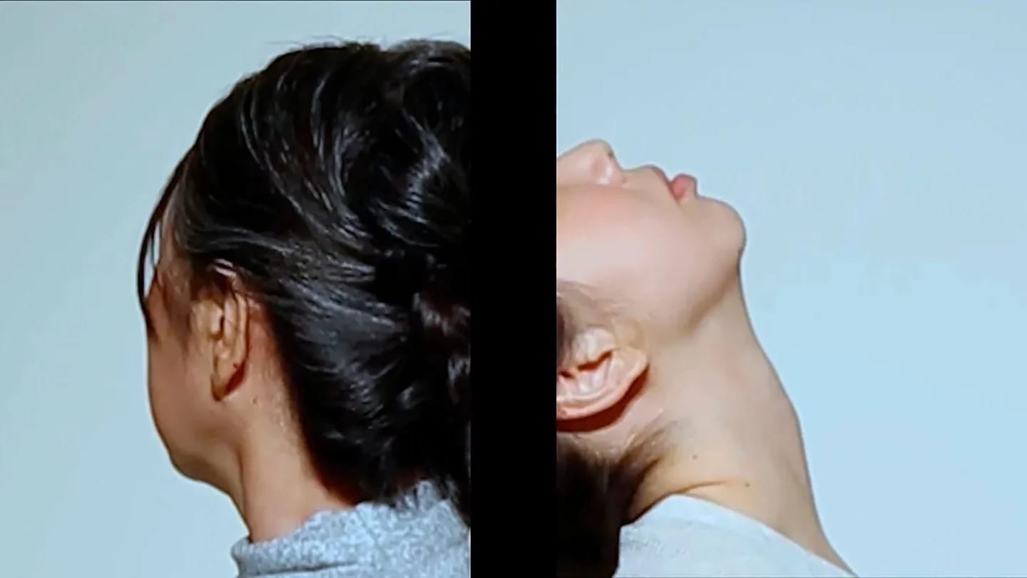 Color still from She is my shadow, I am her echo I by Sunmin Park. Two women appear on separate screens divided by a black vertical border—one turning her head away, the other tilting her head back against a pale blue background.