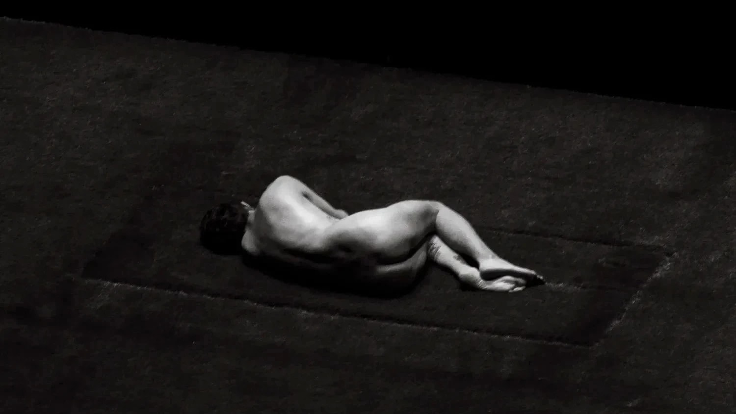 black and white image of naked man from behind in curled position, laying on grass.