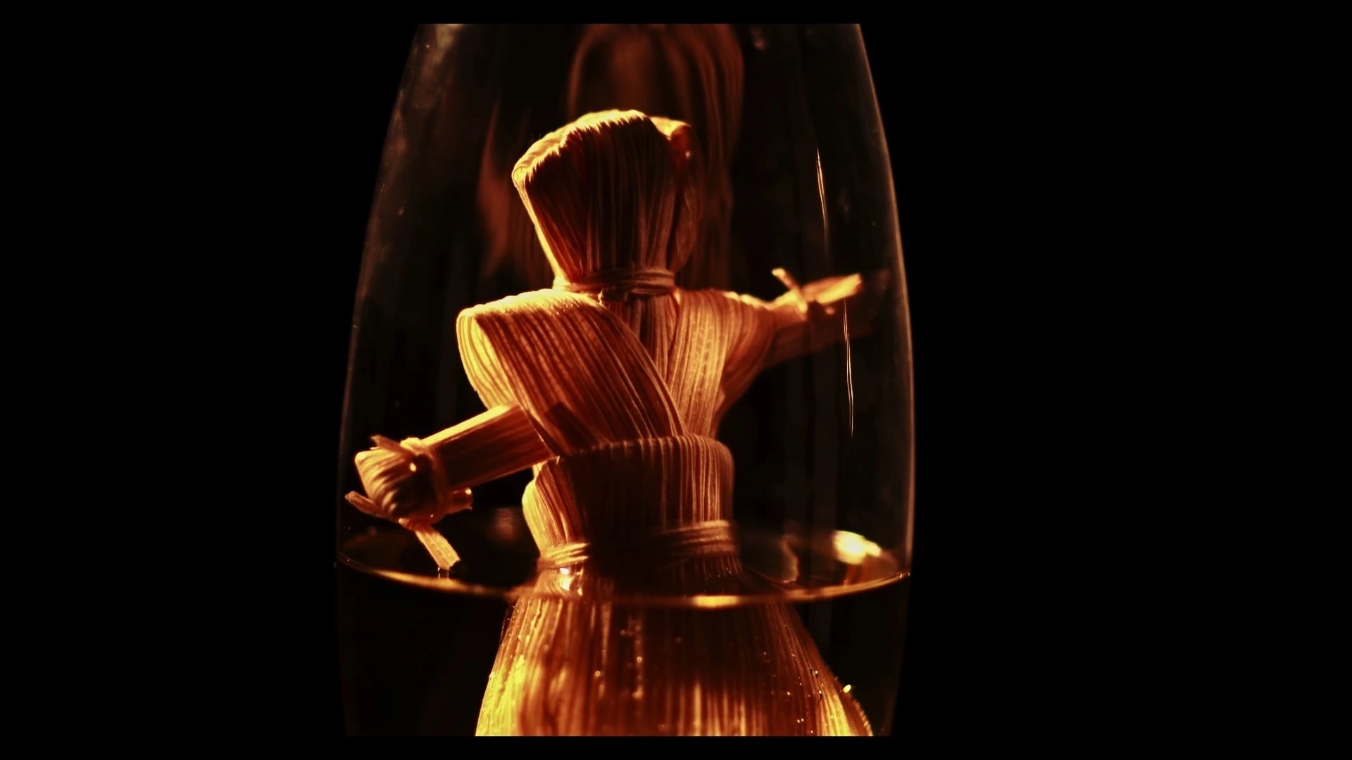 Color video still of a straw doll in yellow liquid encased in glass.
