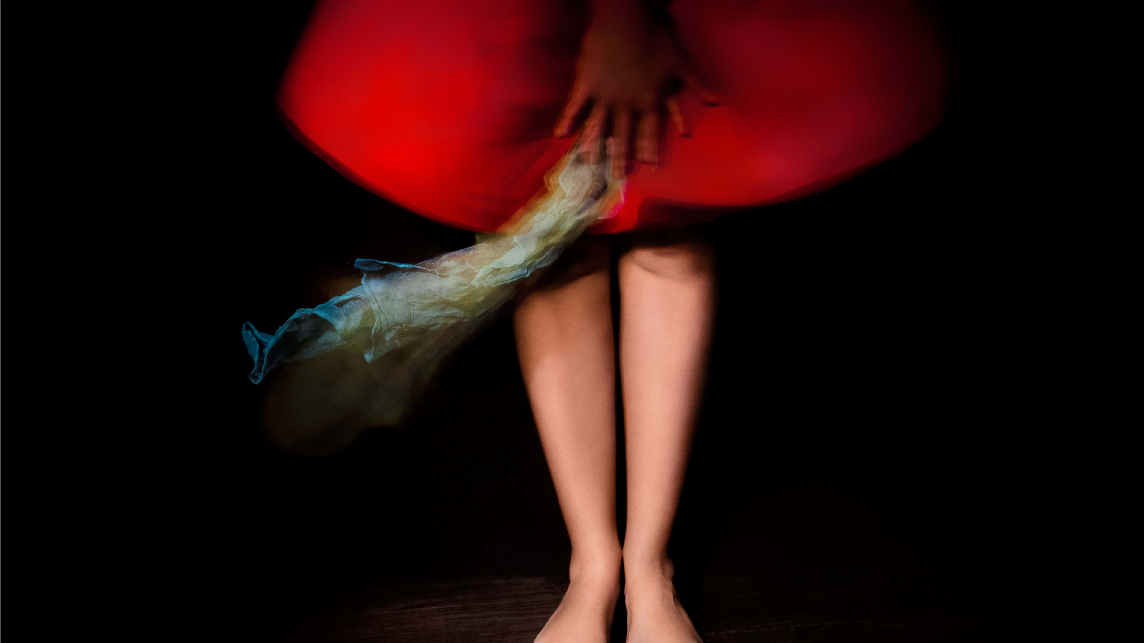 A photograph of a person from the waist down. Their toes are painted with black nail polish. They are wearing a red dress that stops at the knee. Their hands are crossed in front of them, and from their hand is dangling a piece of blue fabric. The dress and fabric are blurred, slightly in motion. The background is black.