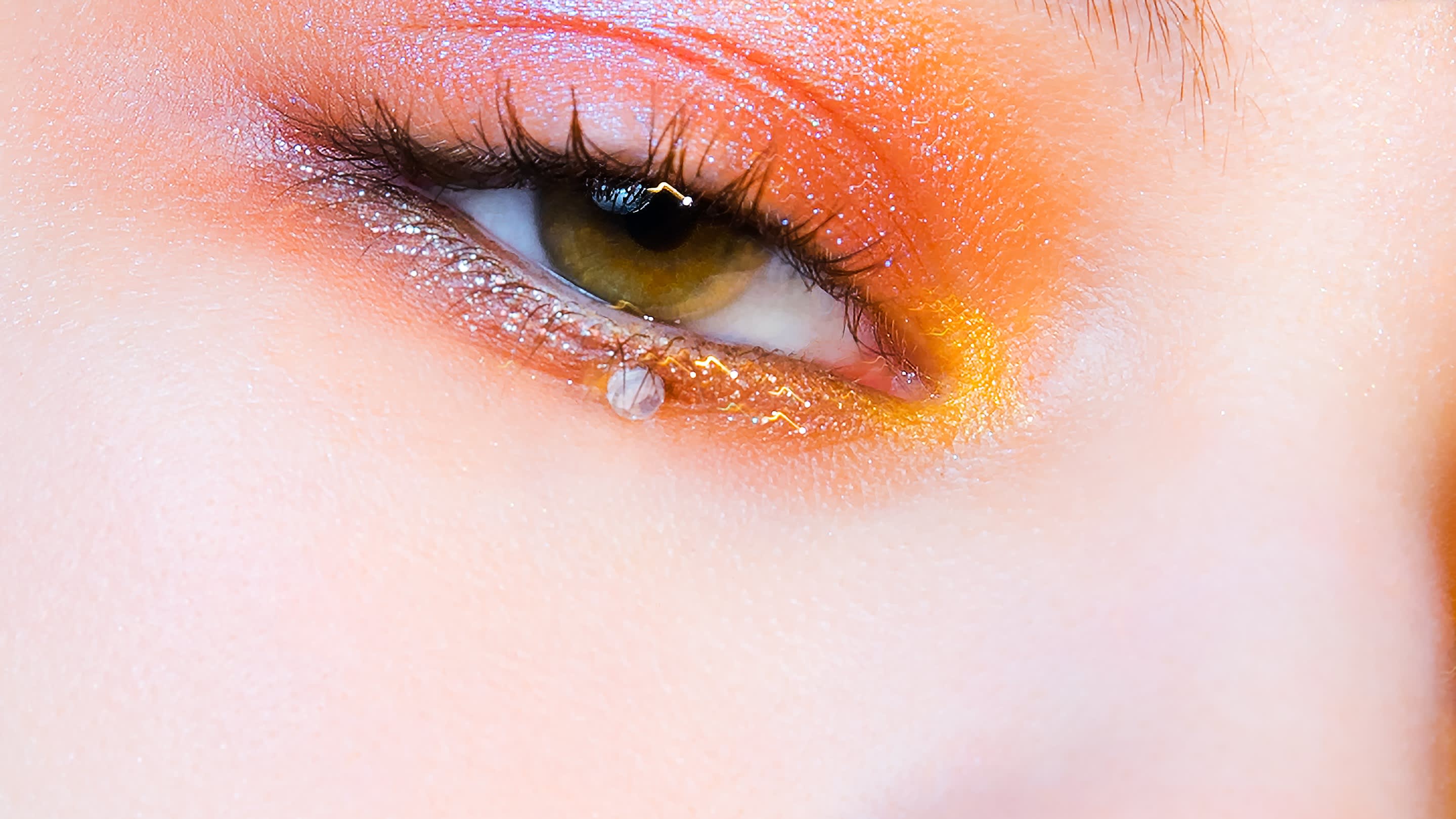 A closeup, studio beauty photograph of a woman’s face. She has white bangs. She is wearing metallic orange and pink eye shadow, light blush, and light pink lipgloss. She has a crystal in the middle of each bottom eyelid. Her hazel eyes are looking down and to the left, off frame.