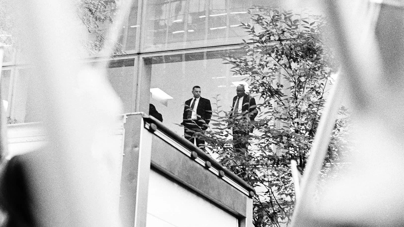 Two men in suits observe a protest from an elevated position