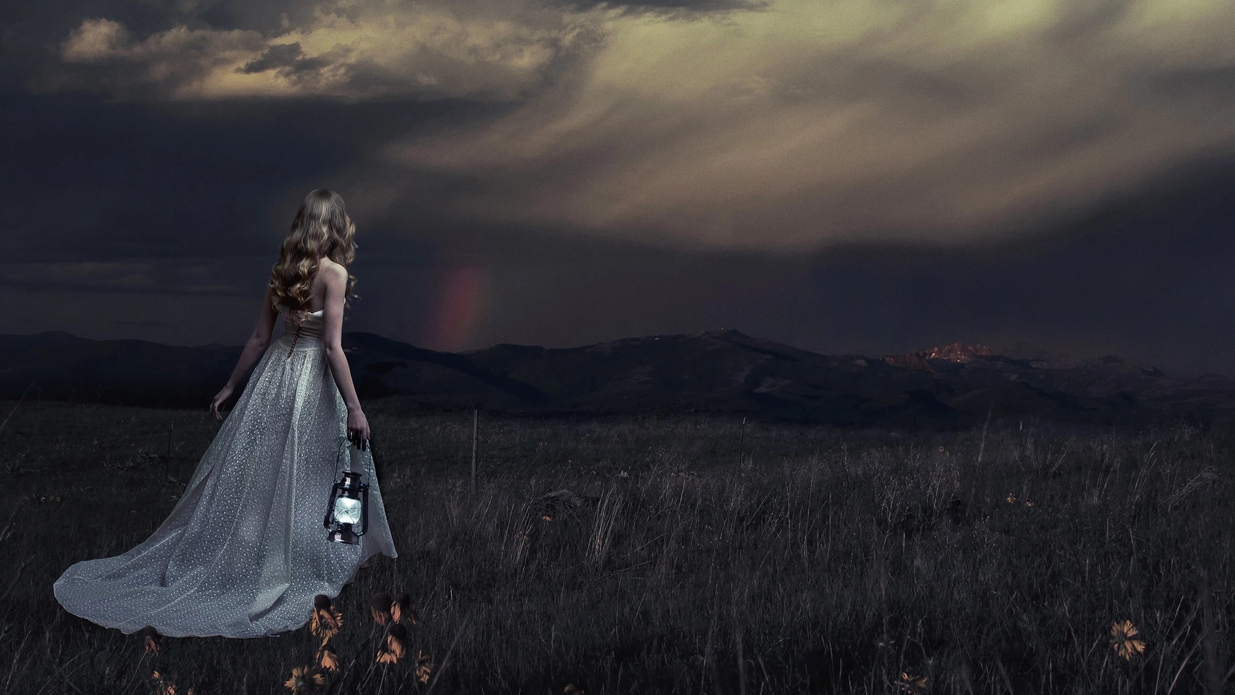 A photograph of a woman in a long pale dress, standing in a dark field and holding a lit lantern.