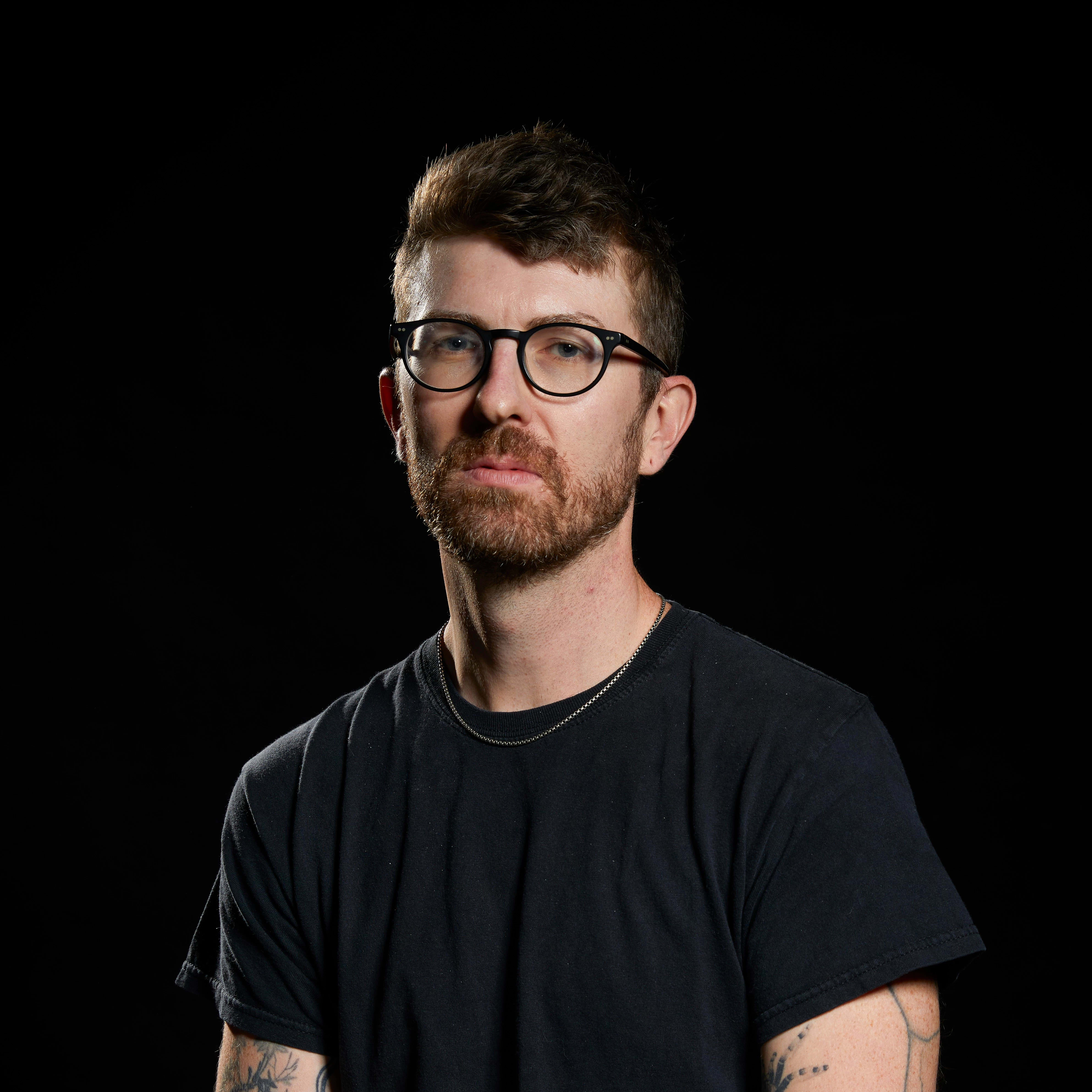 portrait of a man in a studio environment with a dark background wearing a black tshirt. He is wearing glasses and has a silver necklace. 