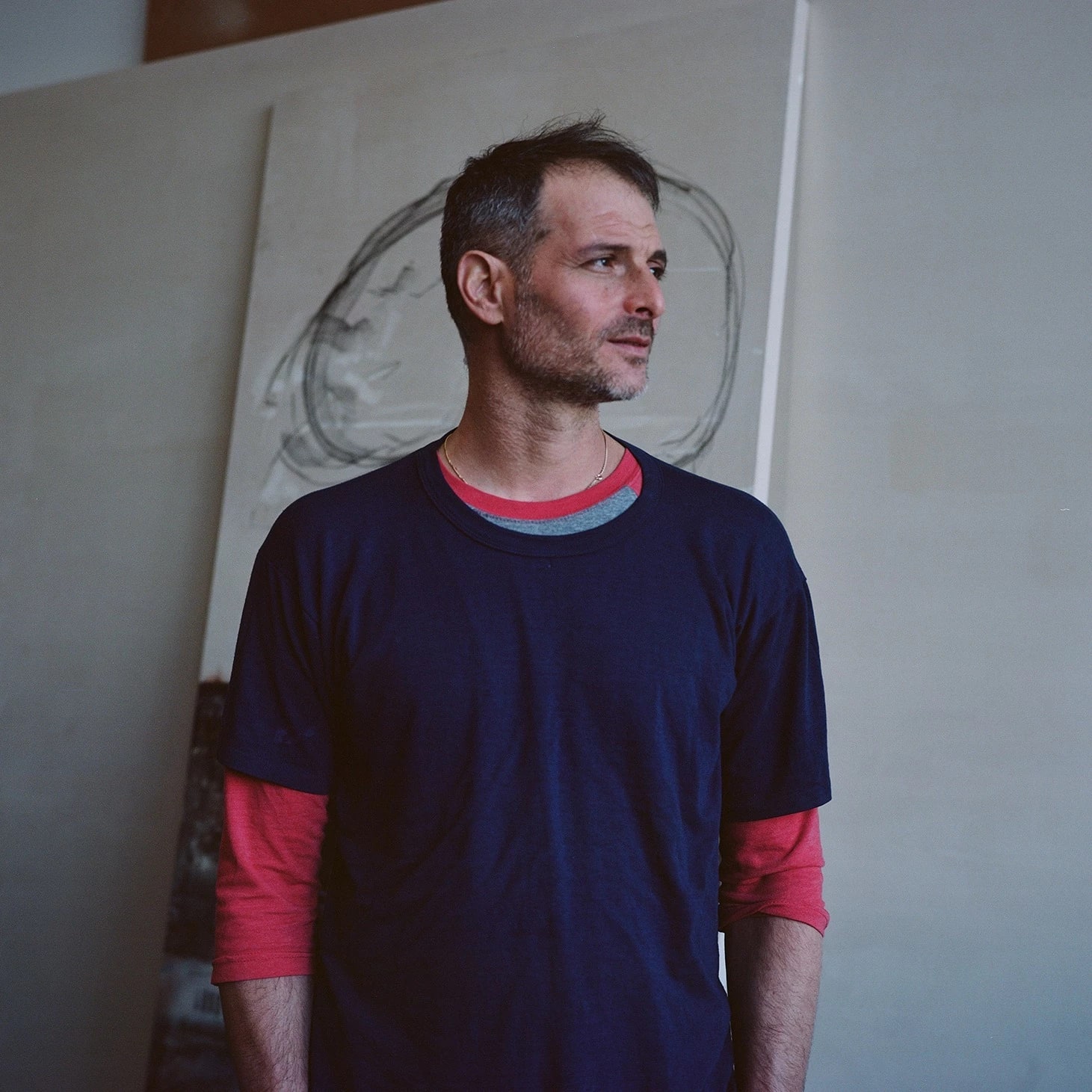 Uri Aran in a navy t-shirt and red long sleeves stands in an art studio, gazing to the side, with a large abstract canvas behind him.