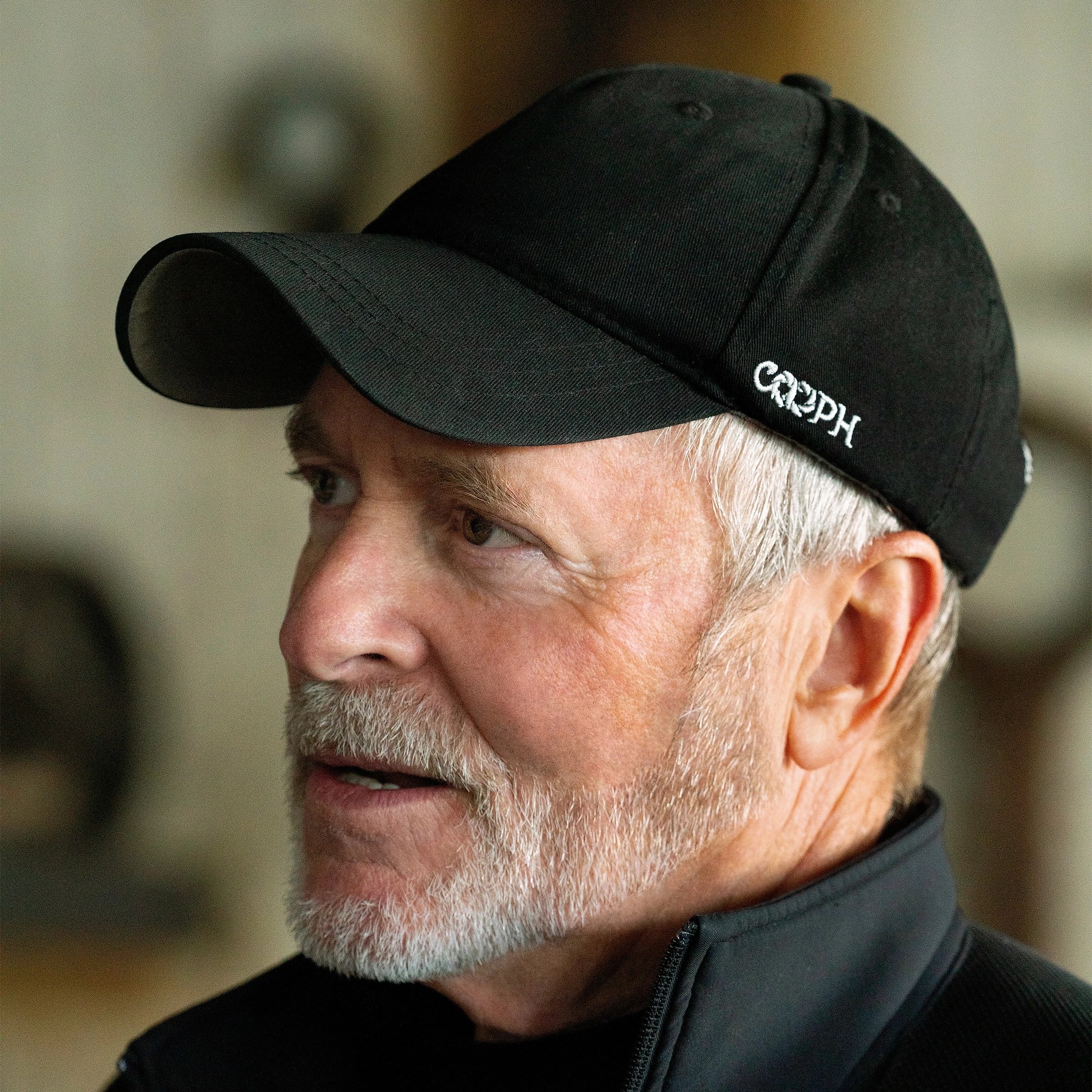 A portrait of Greg Gorman looking to the left of the image. He has silver hair and beard, and is wearing a black baseball cap that says COOPH