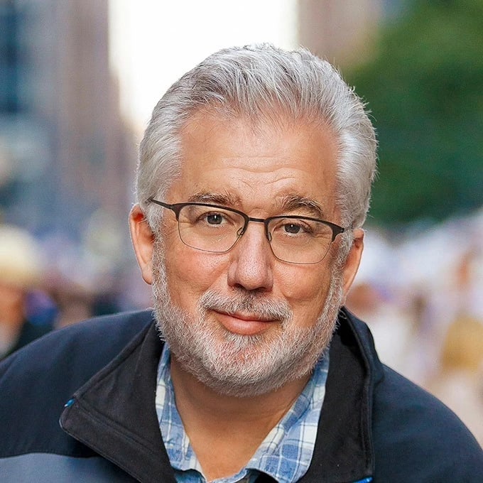 A portrait of Jack Reznicki, who is looking into the camera with a smile. Jack has silver hair, and he is wearing a pair of glasses with a black, thin frame. This photo of Jack is taken on a busy sidewalk.