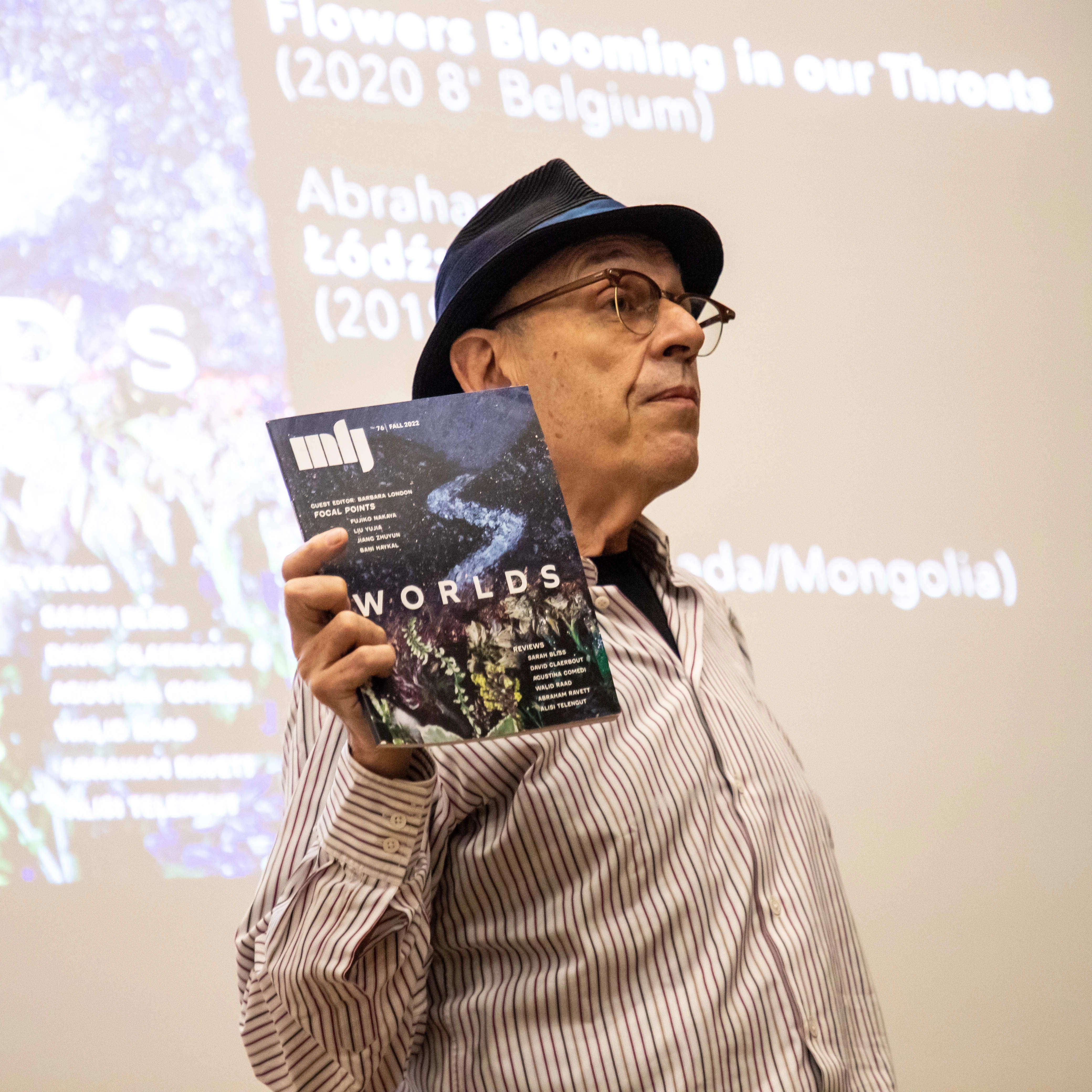 Image of a man wearing a hat, glasses, and a striped button up shirt holding a magazine to an unseen audience.