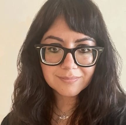 Portrait of Kat Irannejad smiling in the camera, wearing a gold necklace and black framed glasses.