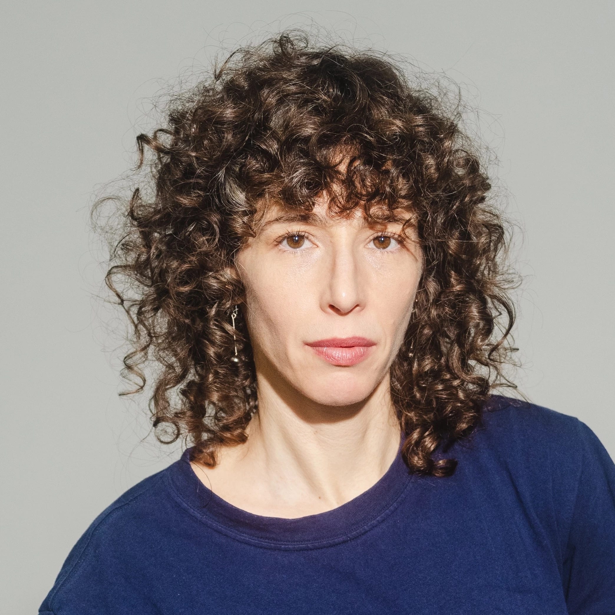 Image of a woman looking into the camera. The woman has curly brown hair and is wearing a blue shirt.