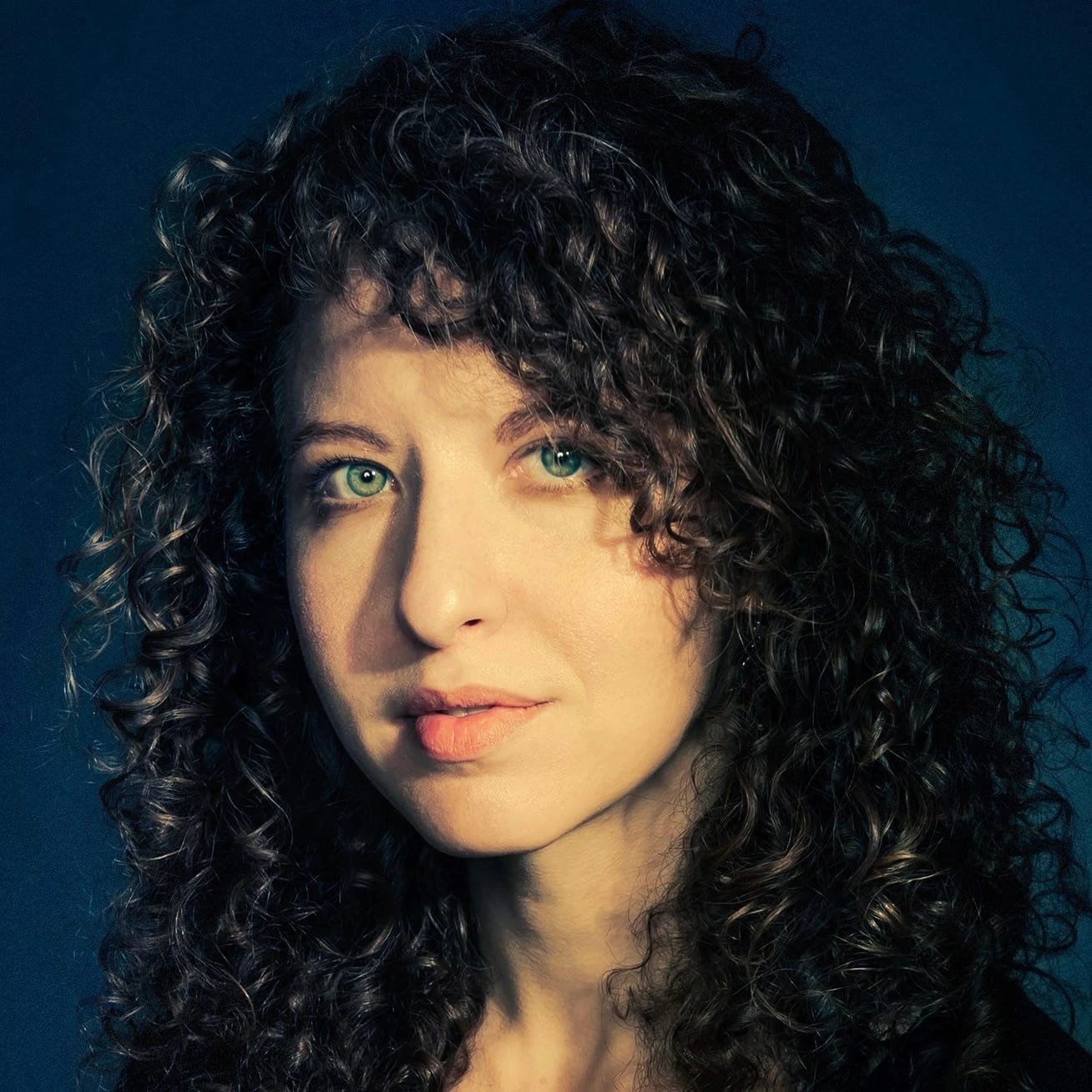 Image of a woman with curly brown hair looking into the camera. There is a blue background.