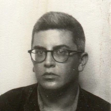 Black and white photo of white person with short graying hair, wearing round glasses, not smiling, looking at camera.