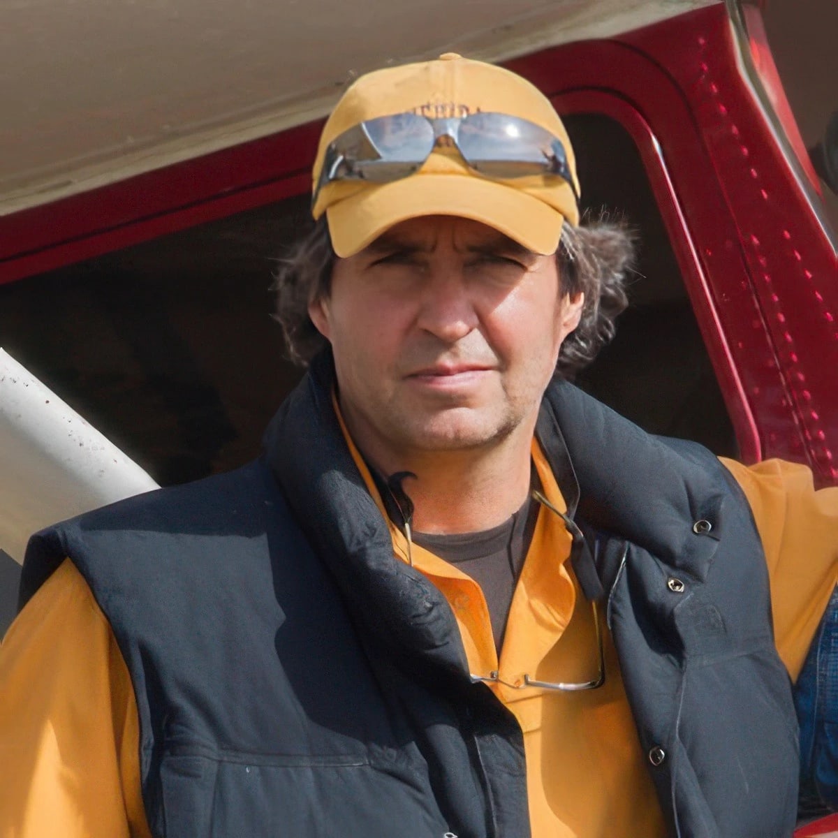 Image of a man looking at the camera. The man is wearing an orange shirt, a black puffy vest, an orange hat with sunglasses on top.