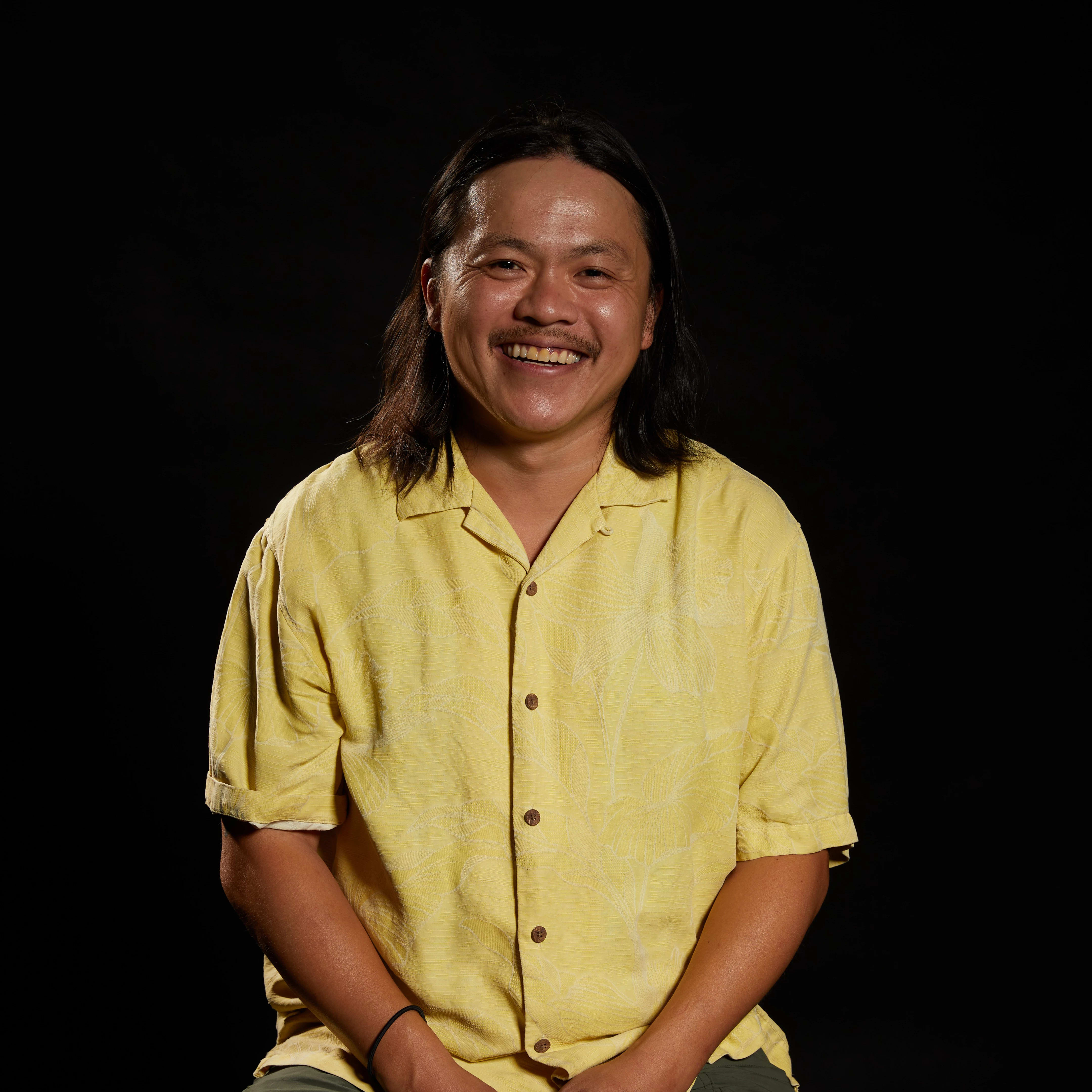 Image of a man smiling at the camera in front of a black background wearing a yellow shirt. The mean has shoulder length brown hair.