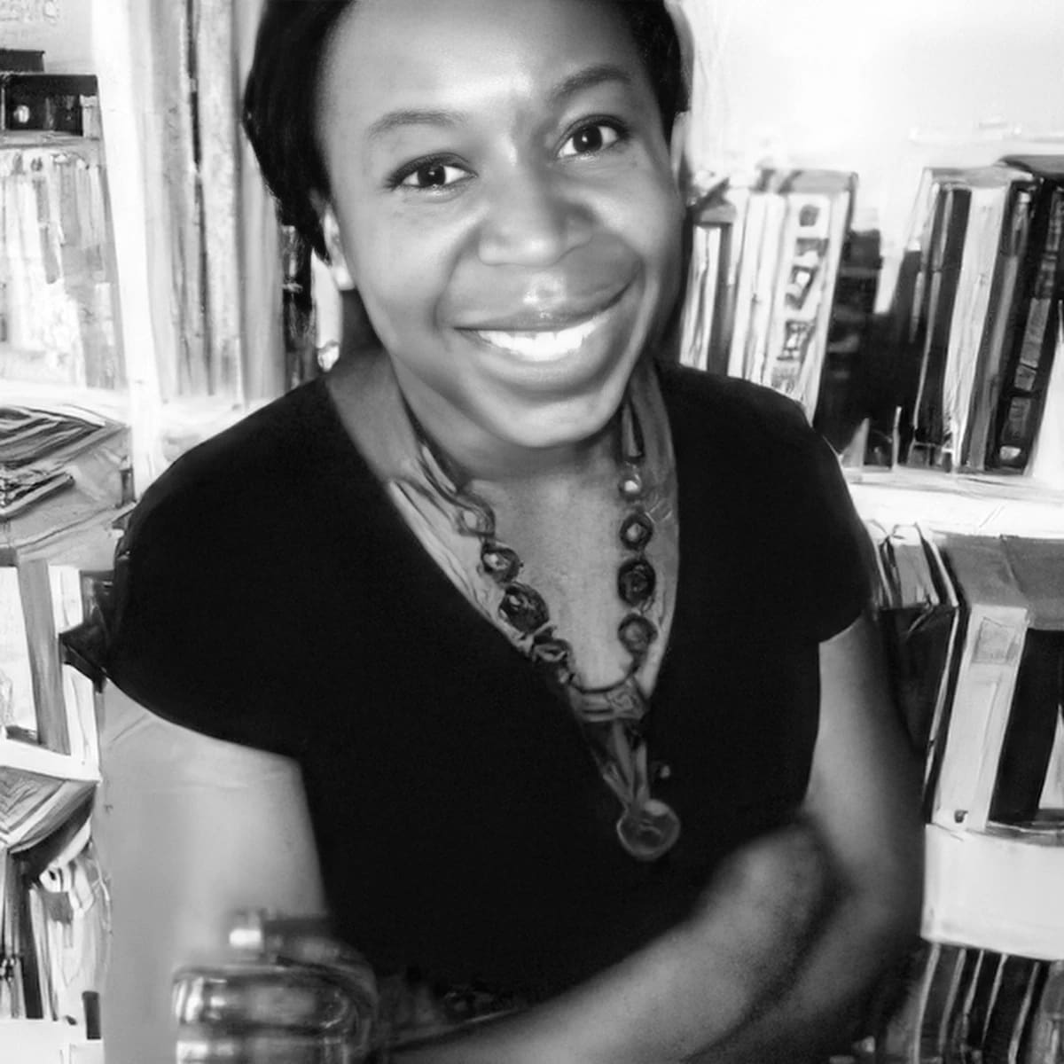 Black and white image of a woman smiling at the camera. The woman has her arms crossed, standing in front of a bookshelf wearing a black shirt and necklace.