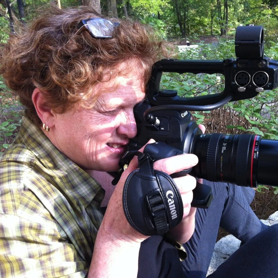 Image of a woman looking through a Canon video camera, shooting video. The woman is sitting on the grass, with curly red/brown hair, glasses on her head, and wearing a plaid shirt.