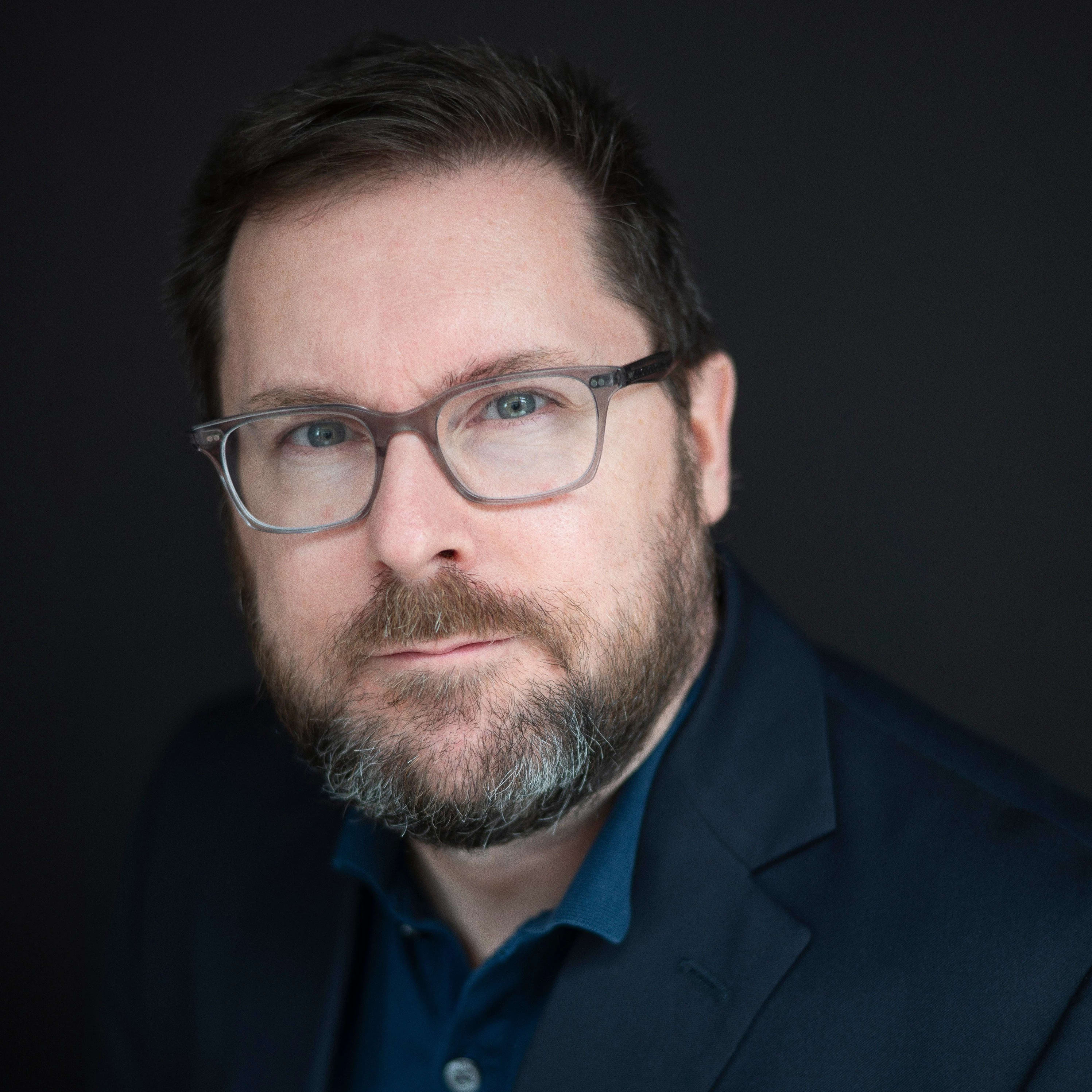 Portrait of Tom Ashe. Tom is wearing glasses and a blue jacket over a blue polo shirt.