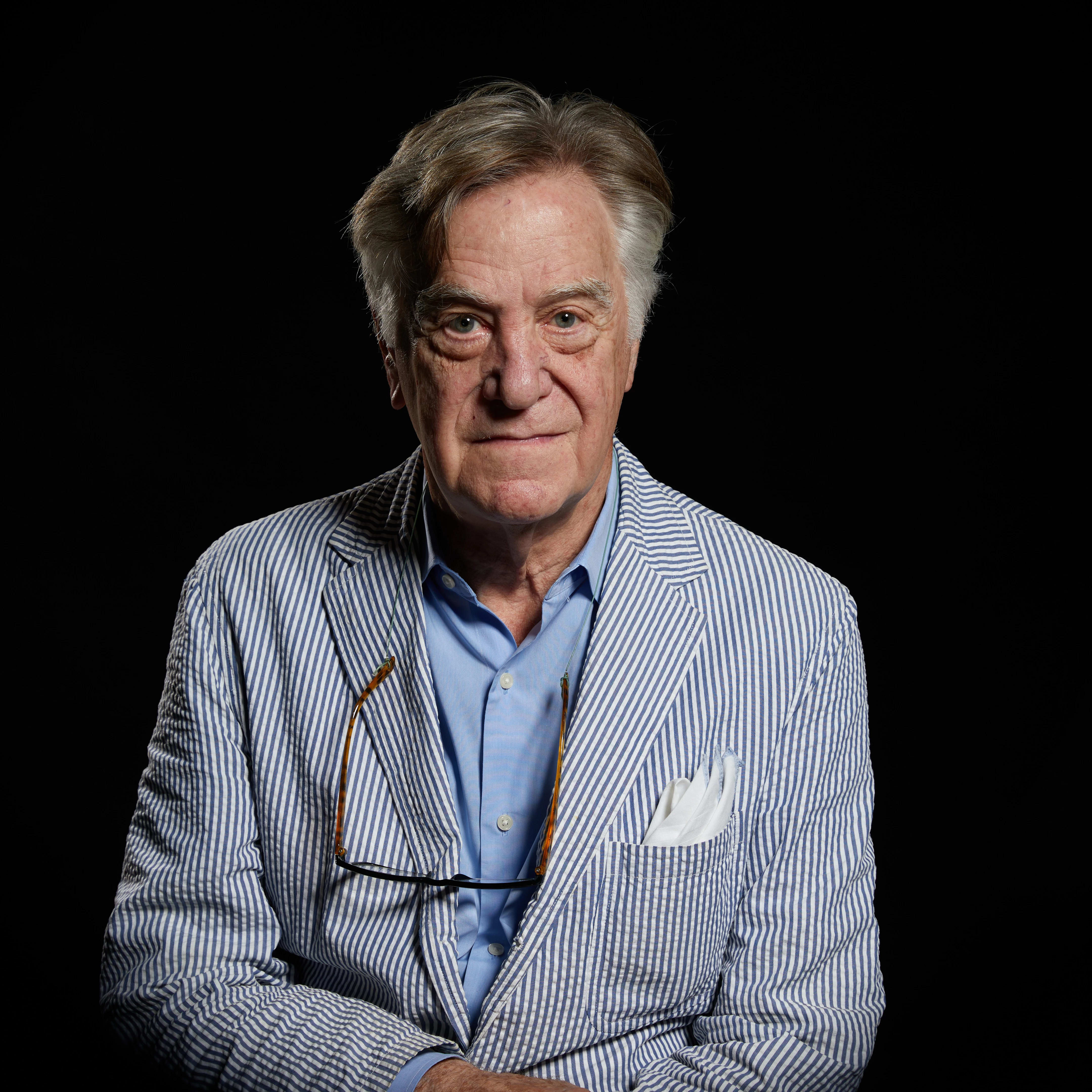 Image of a man in front of a black background. The man is wearing a blue and white pinstripe blazer with a white pocket square and a blue button up shirt underneath; there are glasses hanging round his neck.